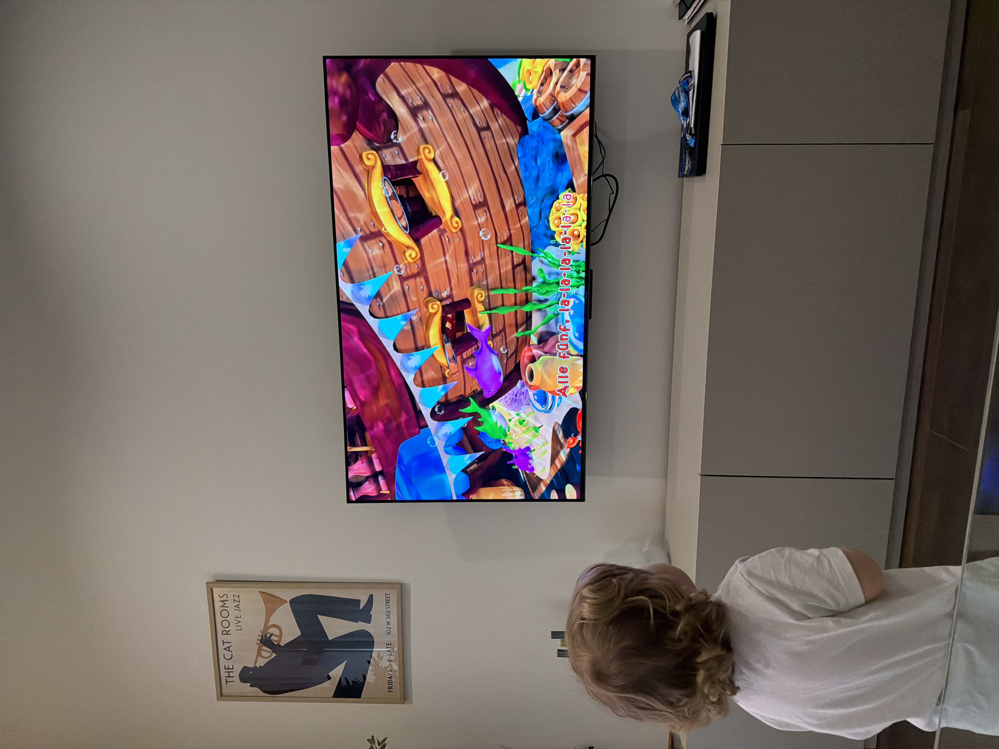 A small child with curly blond hair wearing a white shirt is watching a television mounted on a wall. The television screen displays a brightly colored cartoon scene featuring what appears to be a ship interior. Below the television on the wall is a framed poster advertising "The Cat Rooms Live Jazz," with the location of the event noted as 102 W 3rd Street. The overall environment is a room with light-colored walls, a tall cabinet, and some possible foliage near the poster. There is no mention of Tbilisi, Georgia.