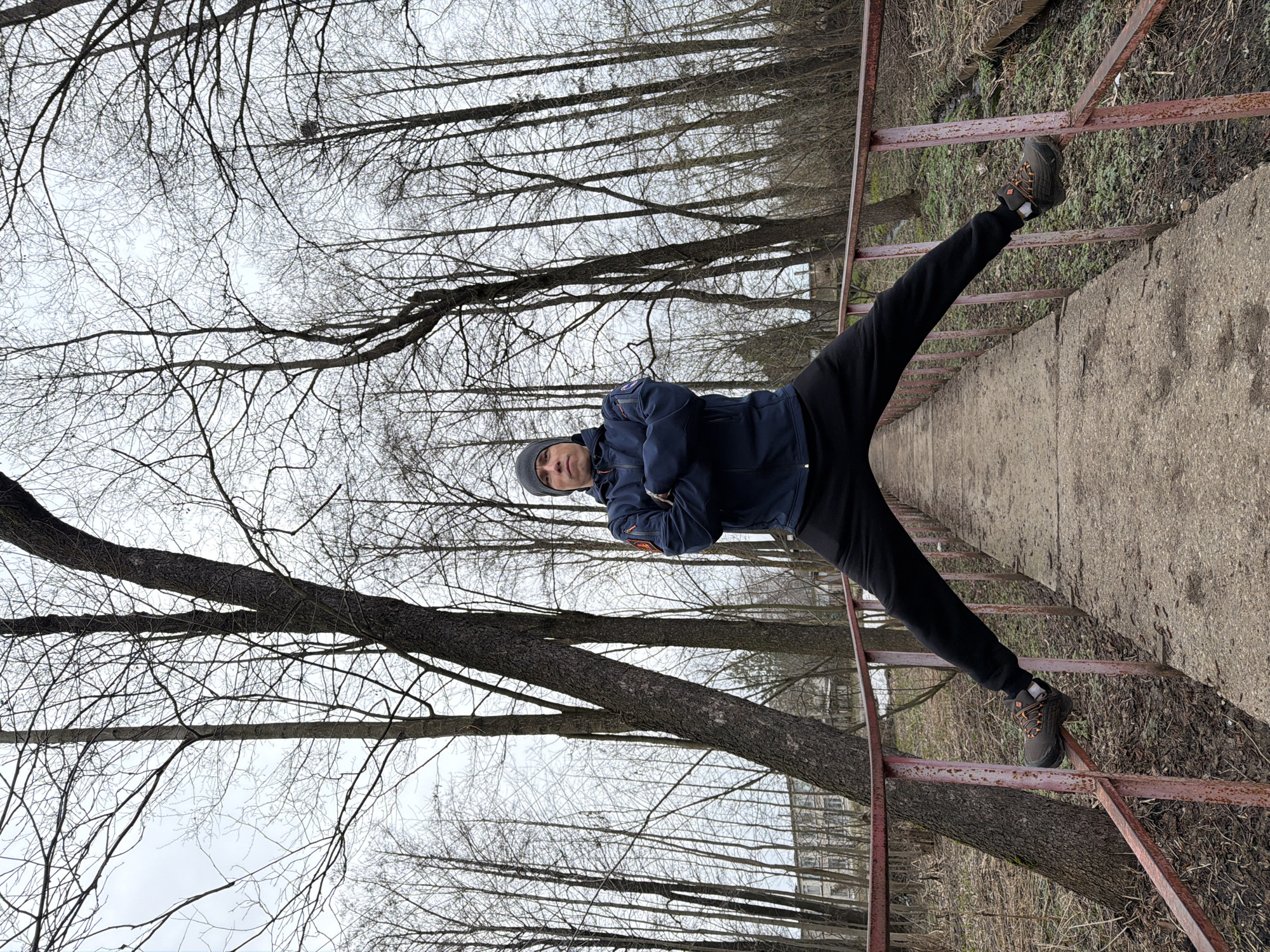 A man in a blue jacket and a gray hat performs a split on a red metal railing in a wooded area in Chaikovsky, Russia. He stands on a concrete path with trees in the background. The sky is overcast.