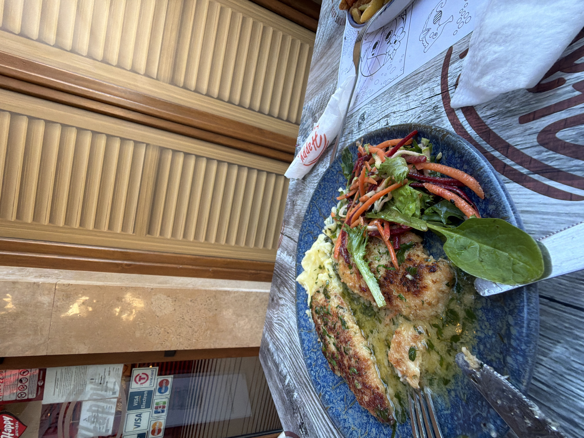 A table at a restaurant in Burgas, Bulgaria, displays a meal consisting of two breaded fish fillets in a light sauce with herbs, mashed potatoes, and a salad of shredded carrots, lettuce, and beets. The meal is served on a blue patterned plate. Utensils, including a knife and fork, are present. A white napkin, folded, rests on a placemat featuring a drawing of a cat and circles. In the background, a wooden door with horizontal slats is visible. The restaurant's entrance, with credit card logos (Visa, Mastercard, Maestro, Electron, Transcard), is to the left. Sunlight reflects off the surface. The overall scene suggests an outdoor dining experience.