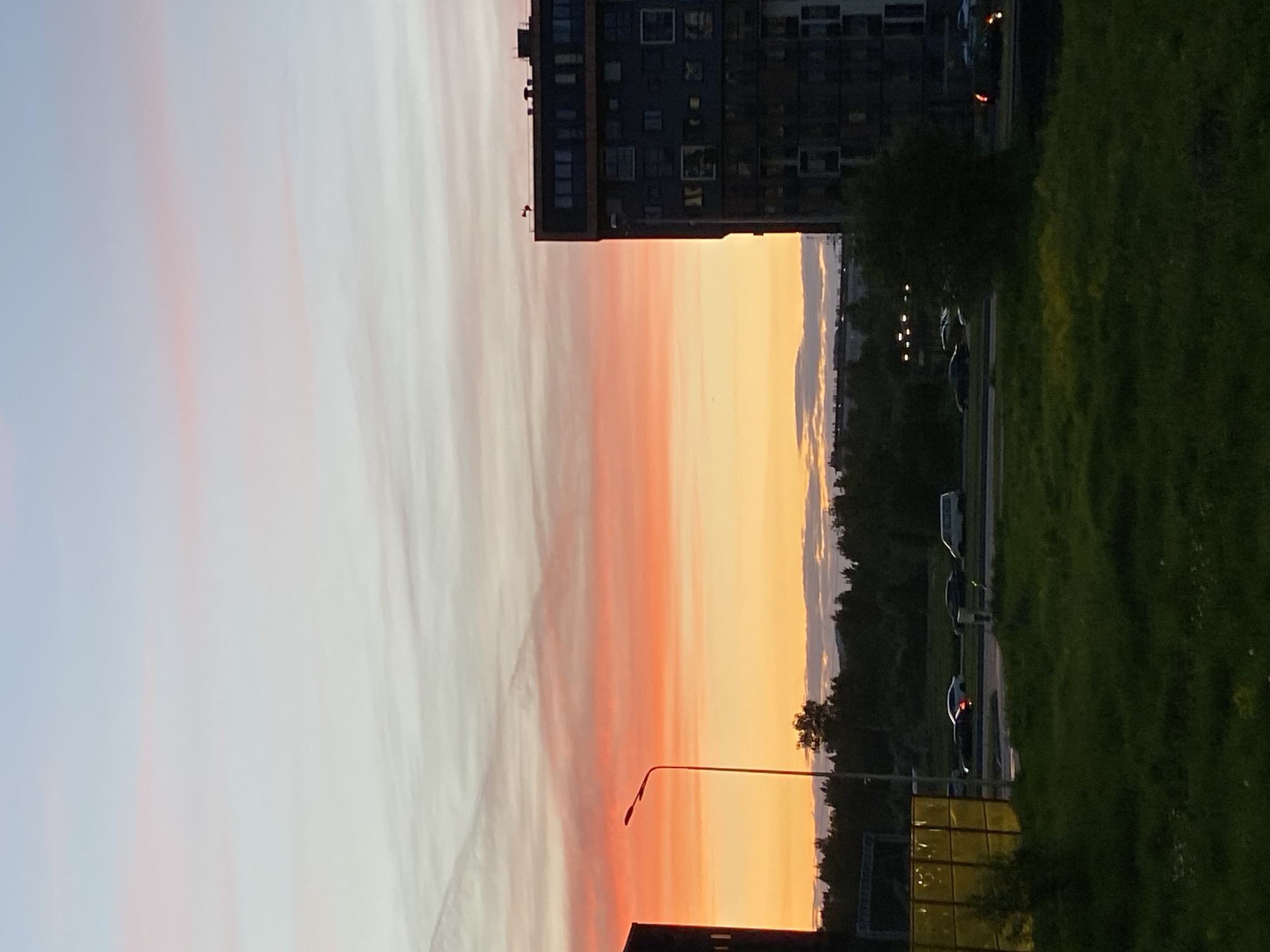 The image shows a building and a grassy field in Saint Petersburg, Russia. There is a tall building with many windows on the top of the frame, and a field of green grass on the right. In between them is a road with cars parked along the side. The sky is a mix of colors, with pink, orange, and yellow hues. There is a streetlight in the lower center of the frame.