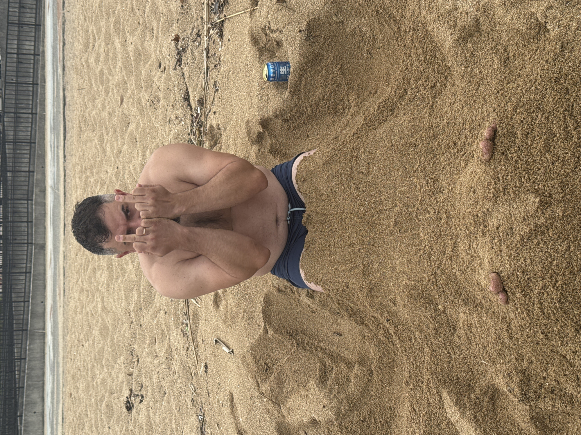 A man is lying on a sandy beach with his feet partially buried in the sand. He is wearing dark swim trunks and is topless. He is extending both hands upward, fingers in the middle finger position. There is a can visible near his waist. A fence is visible in the background. The location is the city of Beppu, Japan.