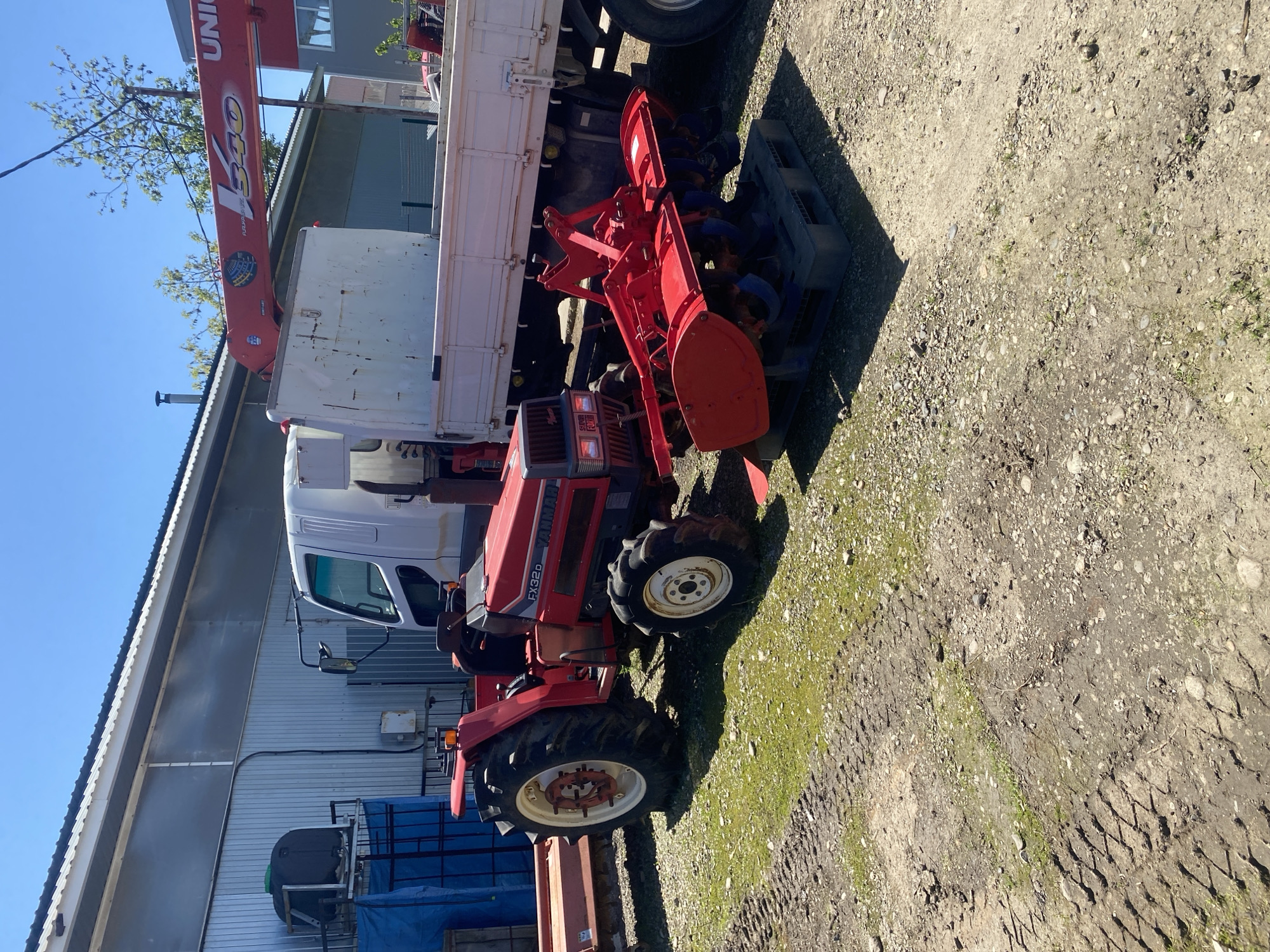 The image shows a red Yanmar FX32D tractor positioned beside a white truck with a red crane arm labeled "UNIC" in what appears to be a rural or industrial setting. The tractor is equipped with a red rotary tiller attachment. The ground is a mix of dirt, gravel, and patches of grass. The truck is parked near a corrugated metal building. There is a blue IBC tote and some wooden planks visible near the bottom of the image. The sky is clear and blue. The location in the city of Neftyanik, Russia, is not explicitly confirmed in the image, so that information is unknown.