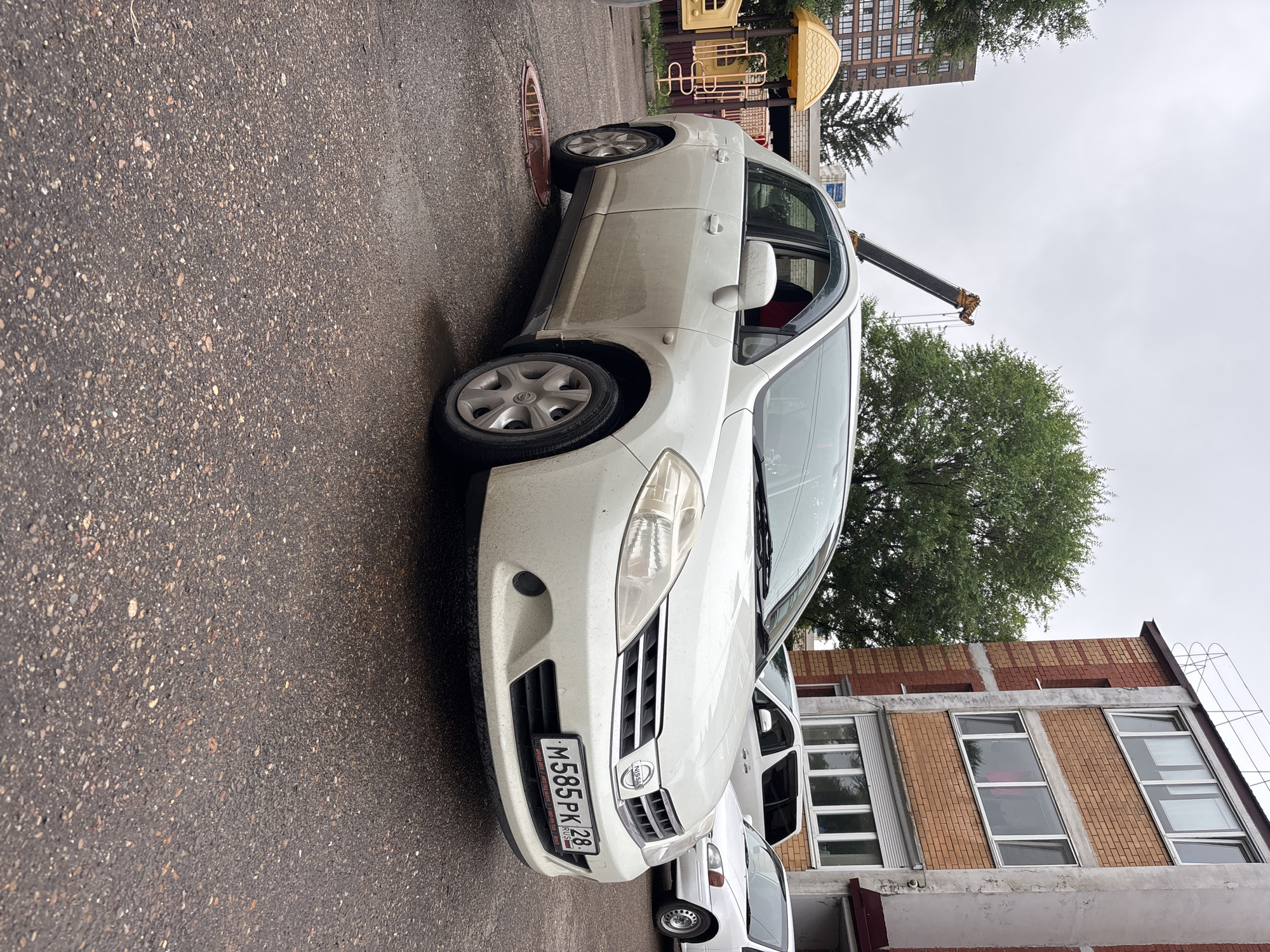 A white Nissan Tiida sedan is parked on wet asphalt in front of a brick apartment building under an overcast sky. The car has a Russian license plate that reads "M585PK 28". To the left of the car is a yellow playground structure. A construction crane is visible in the background. The scene appears to be in a residential area.