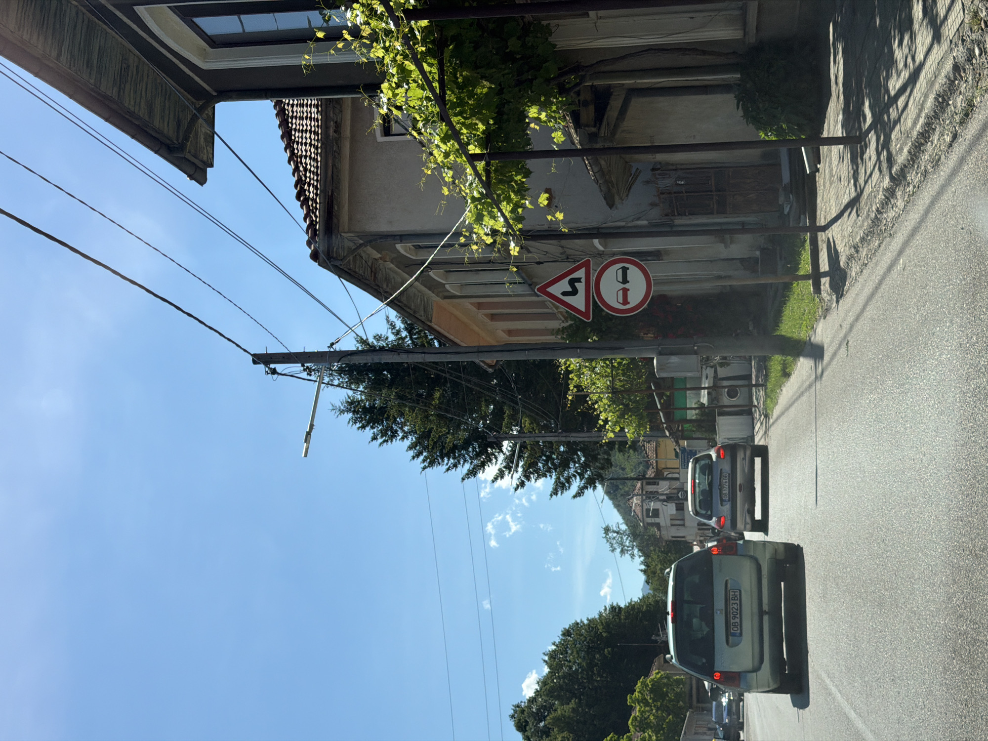 The image depicts a view from inside a vehicle traveling down a street in Beli Osam, Bulgaria, on a bright, sunny day. The road is paved and appears to be a two-lane street. In the foreground, the rear of a light-colored station wagon is visible, with license plate "OB 9023 BH". Ahead of it, another similar car with license plate "OB 0776 BB" is also on the road.
The street is lined with buildings on the right side, characteristic of a European village. One building, in the middle ground, is a light-colored, two-story structure with a tiled roof. A vine with green leaves is growing on the side of this building, partially obscuring it. There are several street signs on a pole near this building. The uppermost sign is a red and white triangular sign with a black wavy line, indicating a winding road. Below it is a circular red and white sign with two black car silhouettes, signaling no overtaking.
On the left side of the street, lush green trees and vegetation are visible, with mountains in the background. Power lines are strung across the sky, supported by utility poles. The sky is clear blue with a few scattered white clouds. The overall impression is of a quiet, rural setting within the village of Beli Osam.