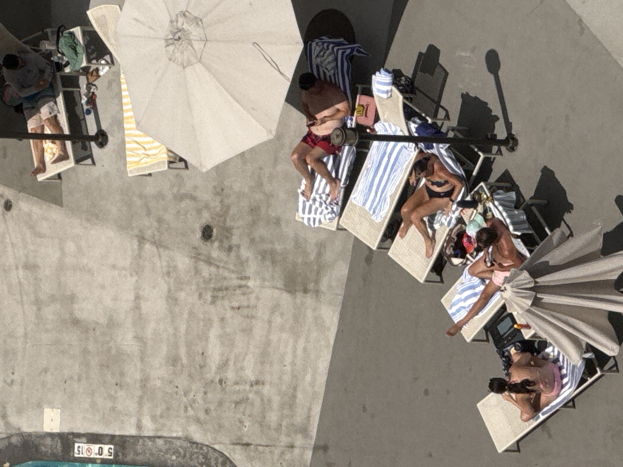 An aerial shot captures a group of people lounging on chairs by a pool on a sunny day. Several individuals are sunbathing on lounge chairs, some under umbrellas. One person is sitting up on a chair, looking at their phone, while others are lying down, presumably relaxing or sleeping. There are beach towels and bags scattered around the lounge chairs. The setting appears to be a resort or hotel with a pool area. The concrete ground and the presence of lounge chairs and umbrellas suggest an outdoor recreational space. The time of day is likely afternoon, given the shadows cast by the umbrellas and people. There are no explicit emotional cues visible from this overhead perspective, but the overall scene conveys a sense of leisure and relaxation. A number "5 0 0 15" is visible on a sign near the edge of the pool, indicating a depth marking. The location is identified as Chicago, USA.
