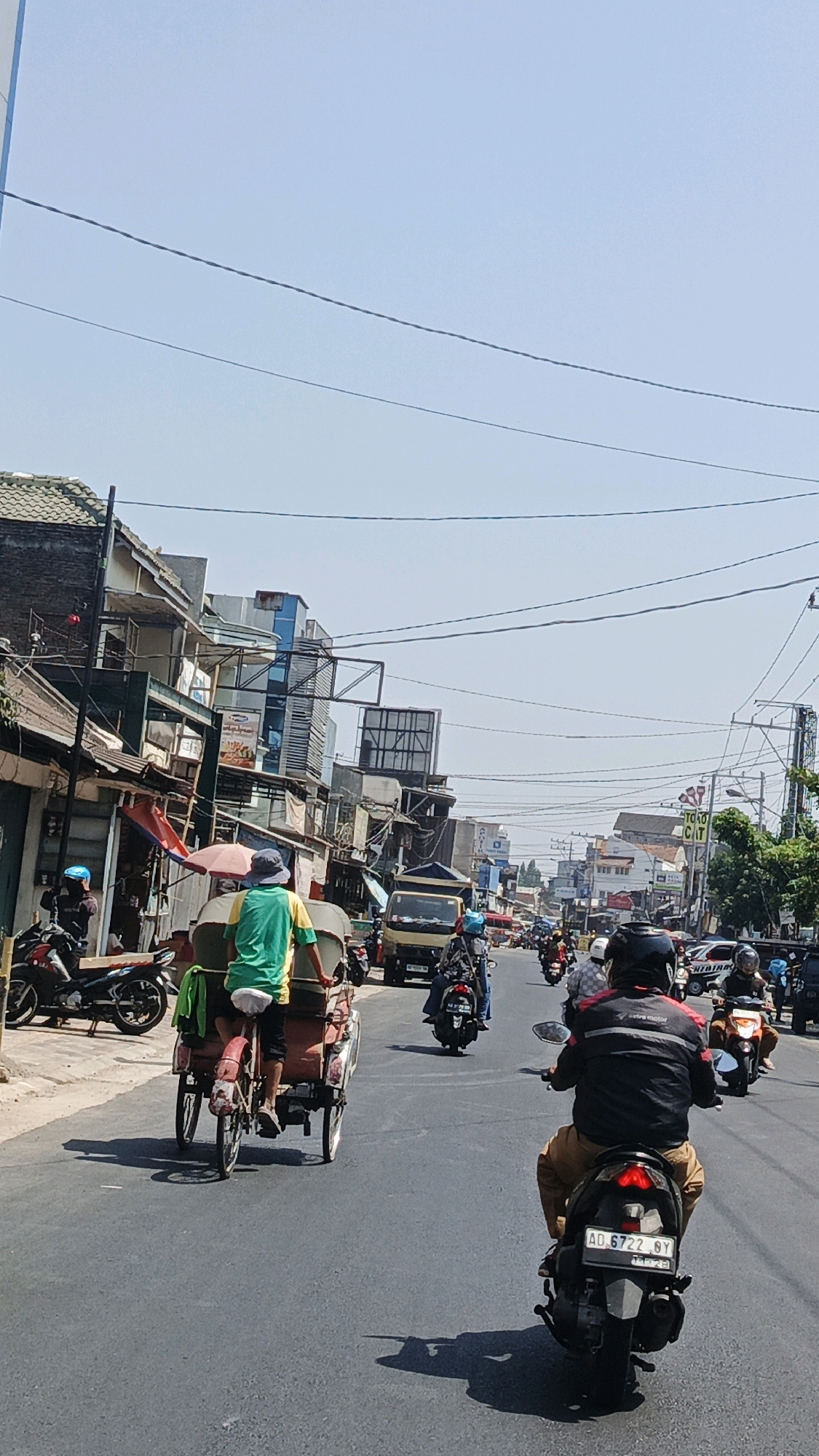 The image depicts a street scene in Surakarta, Indonesia, on a bright, sunny day. The road is paved and busy with traffic, primarily consisting of motorcycles and a few cars and a truck. In the foreground, a pedicab, a three-wheeled vehicle propelled by a rider, is moving down the street. The rider is wearing a green shirt and a hat. Behind the pedicab, several motorcycles are visible, with riders wearing helmets. The buildings lining the street are a mix of commercial establishments, with signs and awnings. Overhead, power lines crisscross the sky. The overall atmosphere is one of a typical urban street scene in Indonesia.