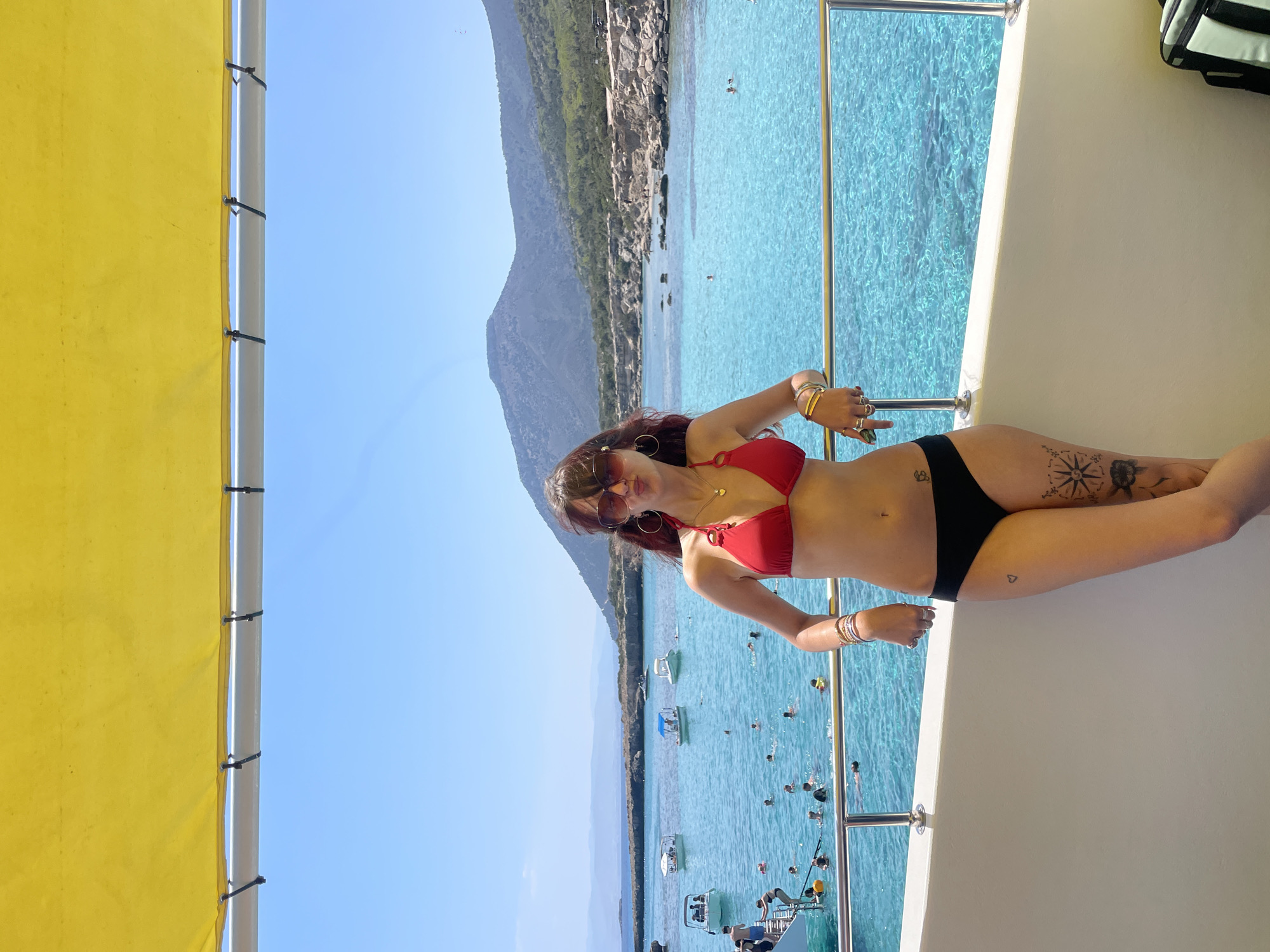 A woman in a red bikini and black bottoms poses on the deck of a boat, leaning against the railing. The boat is on the clear turquoise waters of the Mediterranean Sea, near the coast of Neo Chorio, Cyprus. The coastline features rocky shores and green hills under a bright blue sky. Several other boats and numerous people swimming and snorkeling are visible in the water. A yellow canopy shades a portion of the boat's deck.