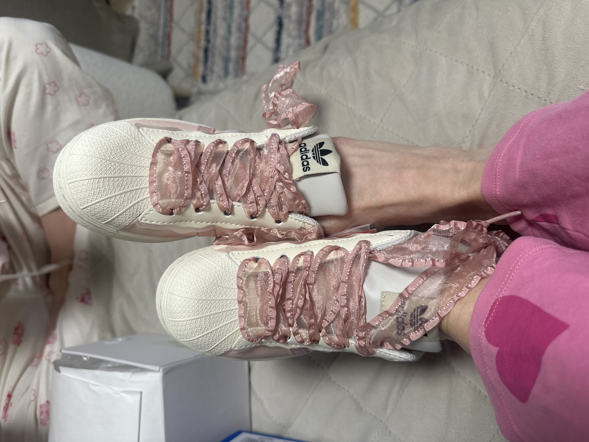 The image shows a pair of white Adidas Superstar sneakers with light pink, ruffled ribbon laces. The sneakers are placed on a quilted light beige surface, likely a bed or sofa. A person wearing light pink pants with a darker pink heart design is positioned on the right side of the image. The backdrop includes soft bedding and a partially visible patterned curtain or wall hanging. A white shoebox is also visible on the lower left side of the image. The location of the image is not discernible from the visual information provided and therefore cannot be determined to be in Votkinsk, Russia.
