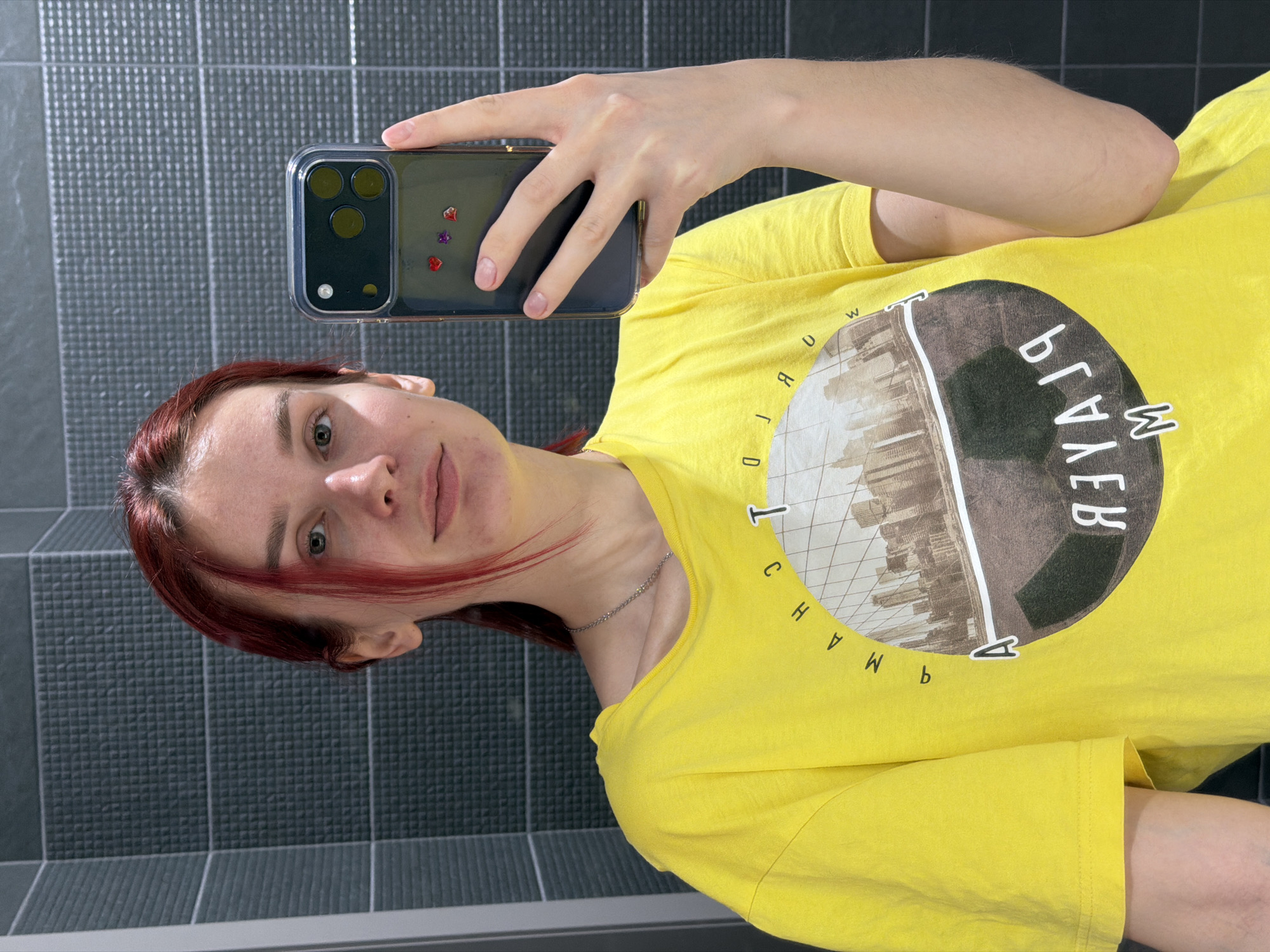 A mirror selfie captures a young person with reddish-brown hair in an indoor setting in Chaikovsky, Russia. The individual, appearing to be female, is positioned with her head on the left side of the frame and her torso extending to the right, due to the image being rotated 90 degrees clockwise. She wears a bright yellow, short-sleeved t-shirt featuring a large circular graphic on the chest. The graphic depicts a silhouette of a cityscape against a lighter background, possibly within a stylized globe design. Visible text on the shirt, reversed due to the mirror reflection, includes "WORLD" (appearing as "DLROW") and "ARCH" (appearing as "HCRA") in a semi-circular arrangement, and "ВГЛЛЕВ" (VGLLEV) on the right side of the circle.
The person holds a smartphone, identifiable as an iPhone Pro model by its distinctive triple camera array, encased in a clear protective cover. Small red and purple heart-shaped or gem-like decorations are visible on the inside of the phone case. Her right hand, with neatly trimmed nails, grips the phone, while her left arm is partially visible. She has light-colored eyes and a neutral to slightly contemplative expression, looking directly into the reflection. A thin silver chain necklace adorns her neck. The background consists of dark gray tiled walls with a textured, possibly dimpled, pattern, typical of a modern bathroom or hallway. No specific time of day or weather is discernible.