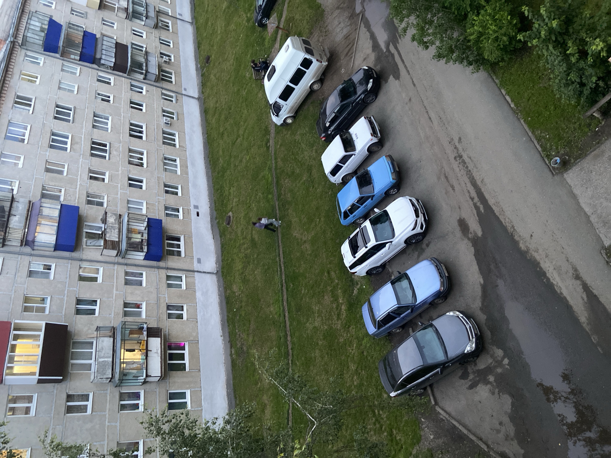 The image depicts a residential building in the city of Beloretsk, Russia. The building is a multi-story apartment complex with light-colored concrete facade. There are several windows and balconies visible. In front of the building, there is a grassy area and a paved parking lot. Several cars are parked in the lot, including a white van, a white SUV, a black sedan, a white hatchback, a blue sedan, and a dark-colored sedan. Two people are walking on the grass in the middle ground. One person is sitting on a bench near the white van. The ground appears damp, suggesting recent rain or puddles. The overall scene suggests a typical urban residential environment.