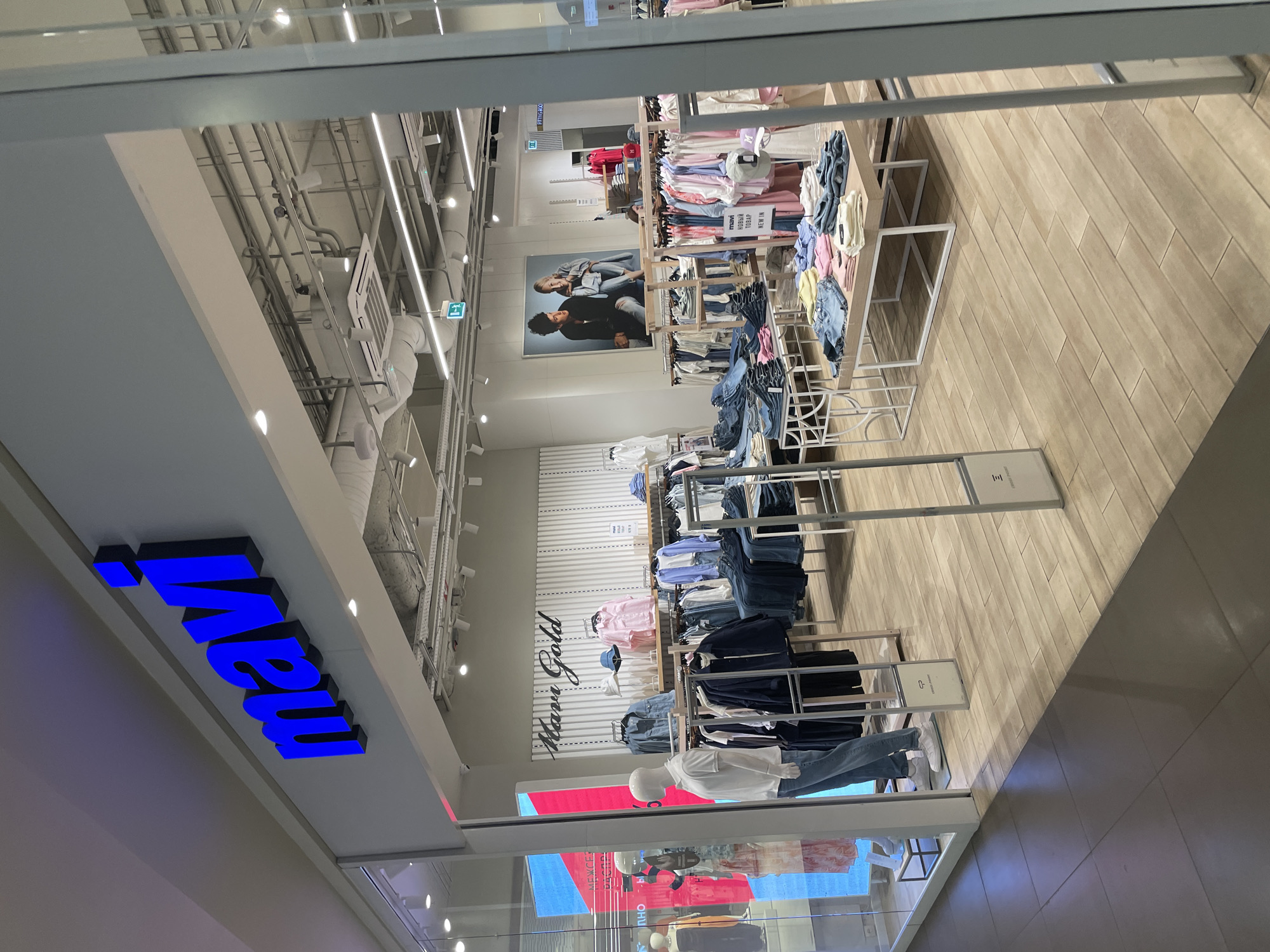 The image shows the interior of a clothing store, presumably a branch of the brand "Mavi," as indicated by the large blue sign. The store is located in what appears to be a shopping mall, given the exposed ceiling with pipes and lighting fixtures. Clothing items, including jeans, shirts, and other apparel, are neatly displayed on metal racks and tables. A mannequin is dressed in a white shirt and jeans near the storefront window. Promotional posters and branding are visible, and the store's name, "Marr Gold," is displayed on a wall. The floor is made of light brown wooden planks. The location may be in a shopping mall in Saint Petersburg, Russia, given the Cyrillic text on the "Новый TОBAP" (New Product) sign and the presence of a "FITTING ROOM" sign in English.
