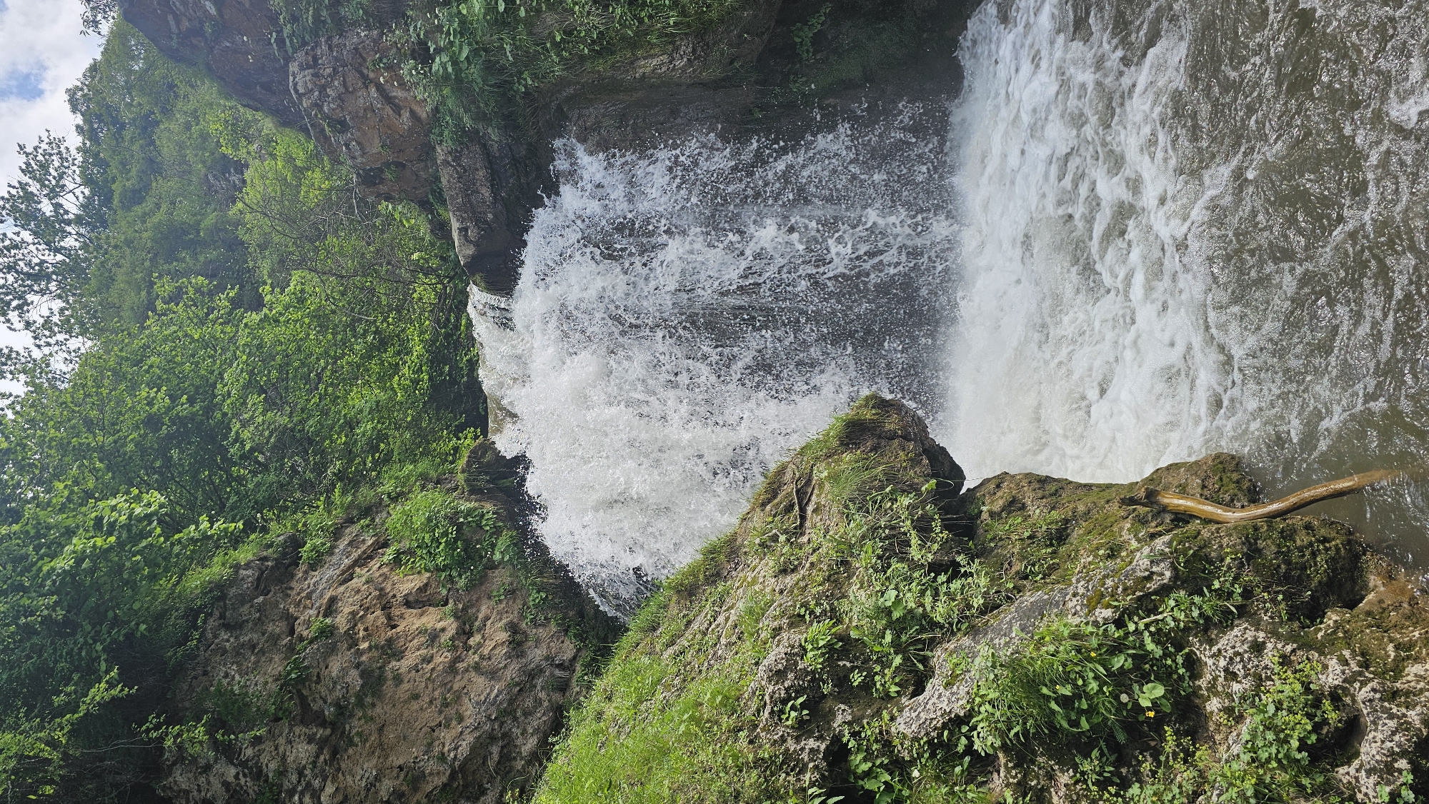 A powerful waterfall cascades over a rocky ledge, surrounded by lush green vegetation and trees. The water is white and frothy as it plunges into the pool below, creating a misty spray. Rocks and soil are visible on the left, with a small branch partially submerged in the water at the base of the waterfall. The sky is cloudy but bright, suggesting daylight. The scene is natural and appears to be in a remote, undeveloped area. It is not possible to determine the specific location in Krasny Kurgan, Russia from the image alone.