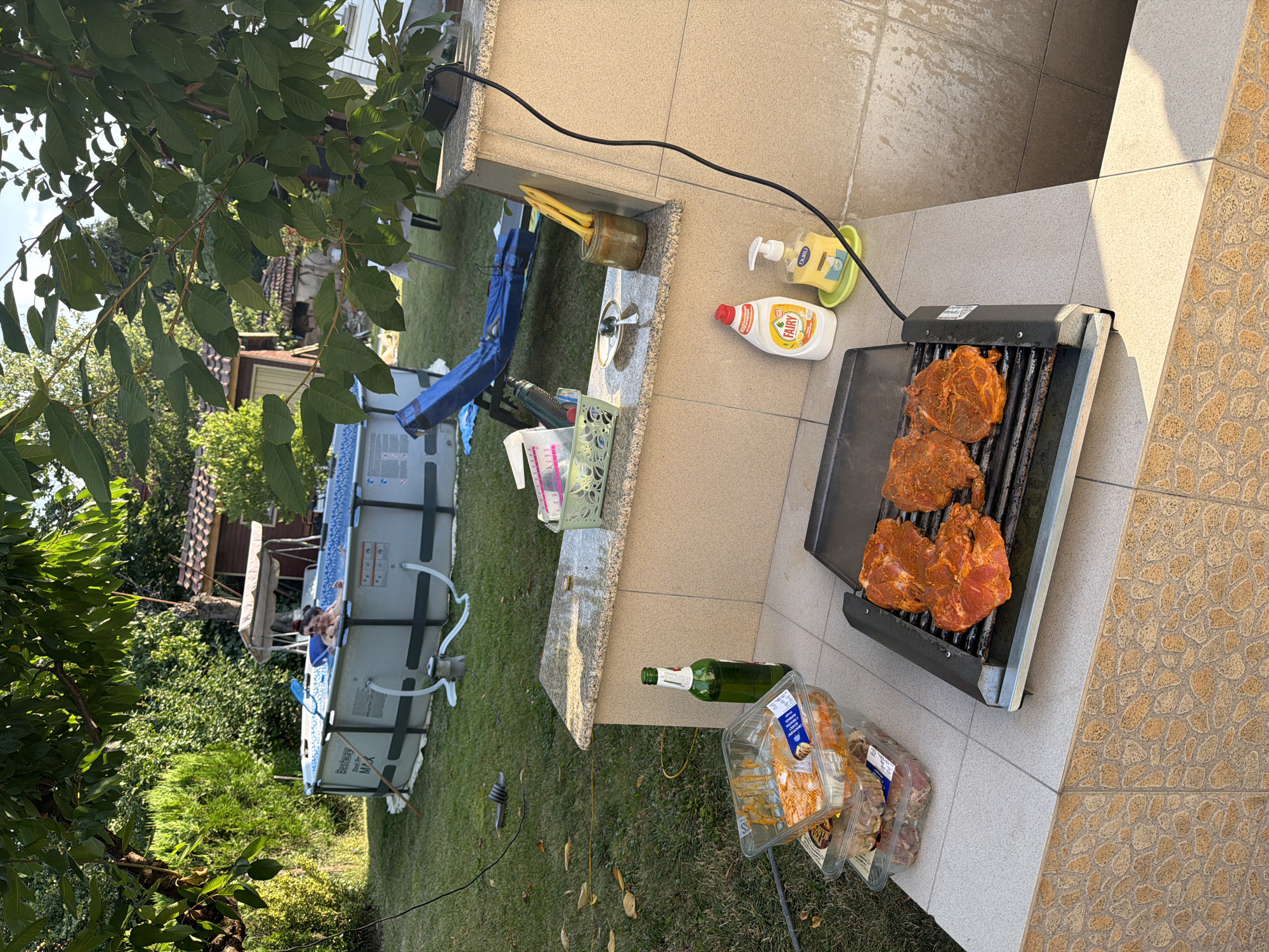 In a backyard setting that appears to be in Bozhurishte, Bulgaria, a barbecue is underway. Raw, marinated meat is arranged on a portable electric grill. Nearby, stacked plastic containers hold more uncooked meat. A green glass bottle of beer stands beside the grill. In the background, an inflatable swimming pool is set up, with two individuals enjoying a dip. Lush green trees and foliage surround the area, suggesting a sunny day. The scene conveys a relaxed, outdoor dining and recreation atmosphere.