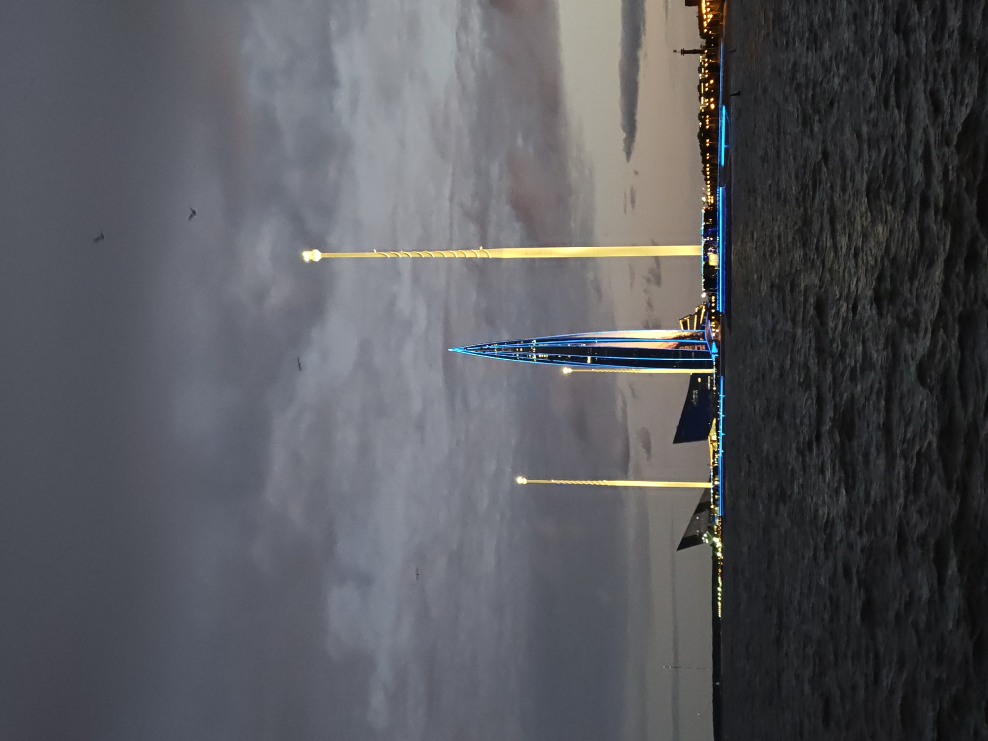 The image depicts the Lakhta Center complex in Saint Petersburg, Russia, at dusk. The iconic skyscraper, characterized by its pointed, twisting design, is illuminated with blue and white lights. Flanking the skyscraper are two tall, slender towers, also lit, creating a striking visual against the darkening sky. In the foreground, a body of water, likely part of the Gulf of Finland, exhibits a choppy surface with reflections of the lights from the shore. The sky is overcast with dramatic clouds, and several birds are visible in flight. The scene conveys a sense of modern architectural grandeur in an urban coastal setting.