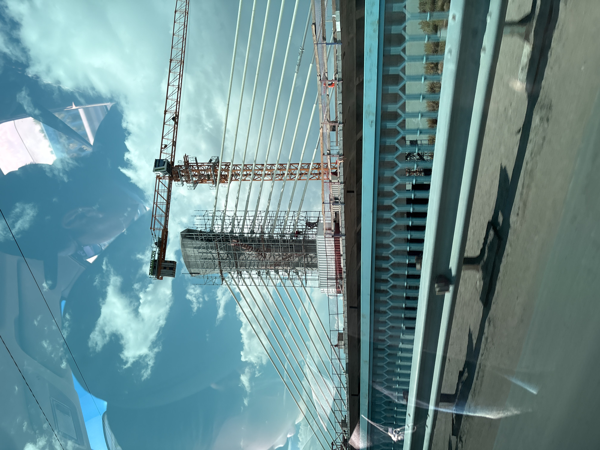 The image shows a cable-stayed bridge under construction, with cranes and scaffolding visible. The bridge's concrete pylon is partially covered in scaffolding, and cables extend from it to the bridge deck. A large orange construction crane is positioned to the right of the pylon, its boom extending across the frame. The sky is cloudy and blue. In the foreground, a blue metal fence and guardrail are visible, with reflections of the sky and clouds on the surface of what appears to be a road or walkway. The location is identified as Ефремовка ауылдық әкімдігі, Kazakhstan.