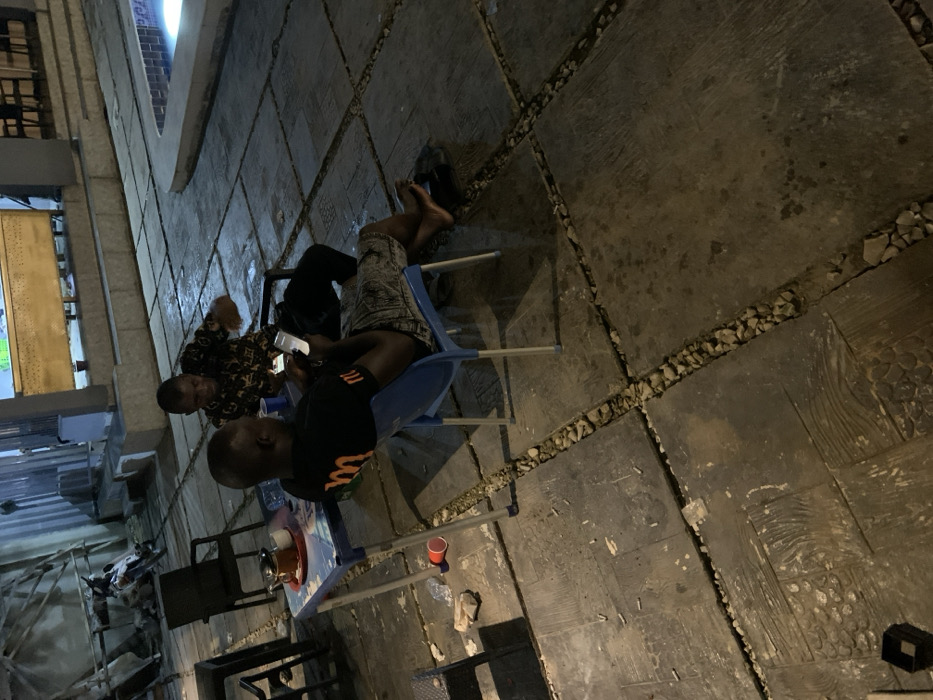 The scene depicts an outdoor patio area at night in Akwanga, Nigeria. Two adult males are seated at a small, light-colored plastic table on a paved surface. The ground is composed of large, dark concrete or stone slabs separated by gravel-filled expansion joints, exhibiting reflective wetness in areas. Artificial lighting illuminates the environment.
In the foreground, a dark-skinned male, with his back to the camera, wears a black t-shirt with an orange "M" logo and patterned shorts. He is seated on a blue plastic chair, barefoot, with his feet resting on the ground, and is focused on a smartphone held in both hands.
Opposite him, a second dark-skinned male, wearing a dark patterned shirt, sits on a black chair. He is also holding a smartphone in his left hand and appears to be gesturing with his right hand.
On the table between the individuals, a dark container with a handle (possibly a teapot), two small white cups on a red tray, and a blue plastic cup are visible. On the ground near the table, a red plastic cup, a crumpled white object, and multiple small white cylindrical objects (consistent with cigarette butts) are present.
In the background, to the left, a white motorcycle is partially visible behind an empty black chair. To the right, an illuminated swimming pool with a blue glow is present. Building structures, including a corrugated metal wall and a raised platform, are also visible in the background.