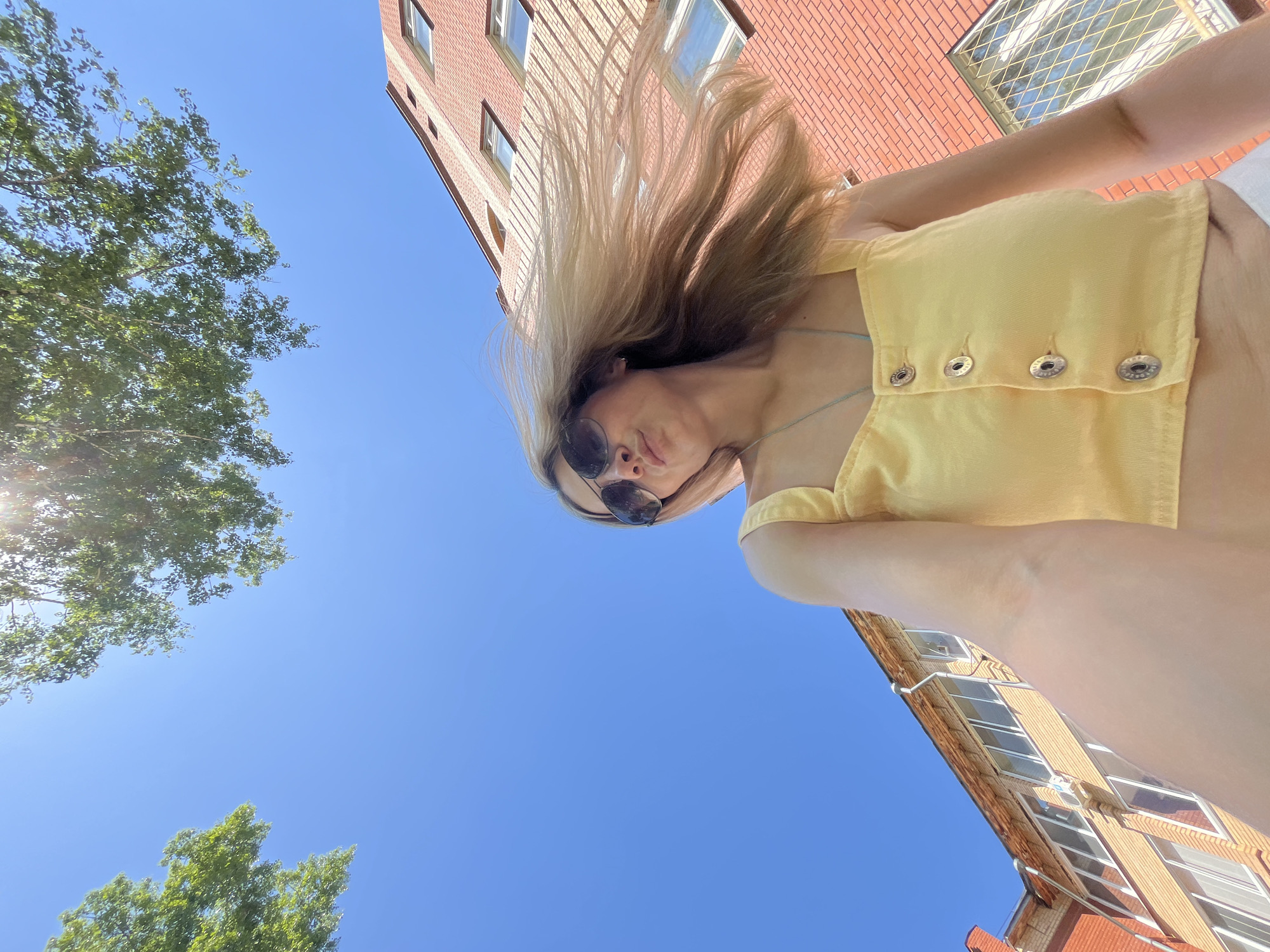 A low-angle shot shows a woman with long blonde hair, wearing sunglasses and a yellow crop top, in an outdoor setting. The sky is clear blue. Behind her are brick buildings and treetops. It is difficult to confirm the location as Chaikovsky, Russia, without further information.