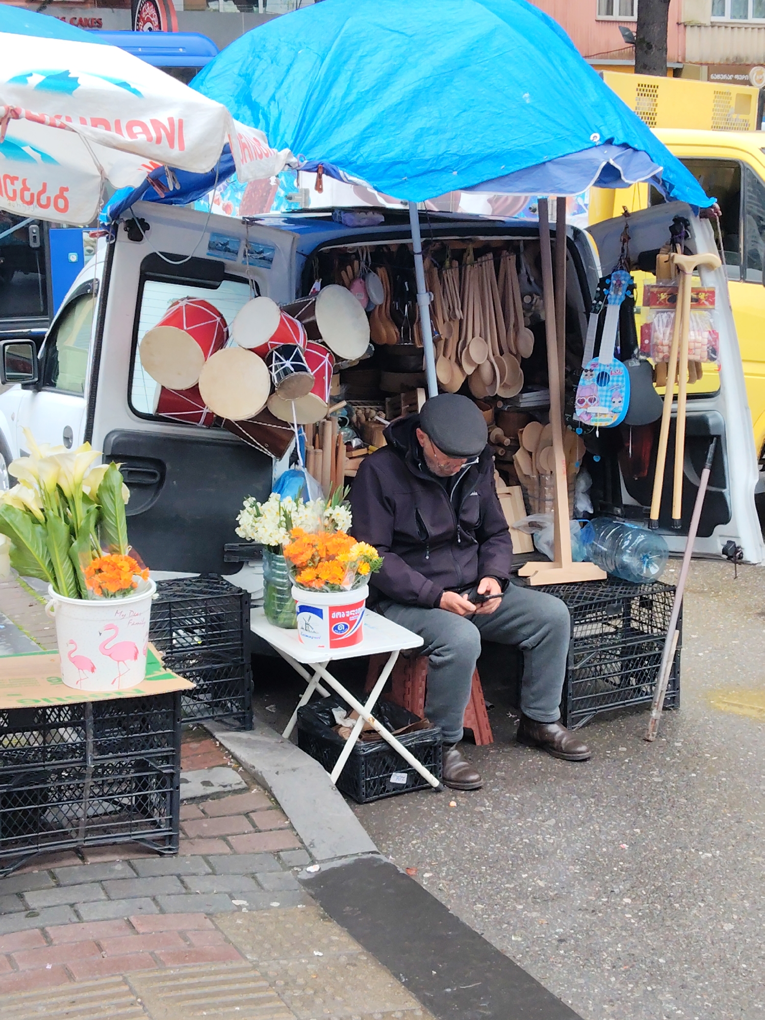 A street vendor sits on a black crate at an outdoor market in Batumi, Georgia. The vendor, a middle-aged man, wears a dark jacket and a gray cap while looking at his mobile phone. Behind him, his goods are displayed in the open back of a white van under a blue tarp. Items for sale include wooden spoons, walking sticks, small toy guitars, and drums hanging overhead. Vases of flowers are on a small table to his left. The street is wet and made of stone blocks, leading to a concrete sidewalk.
