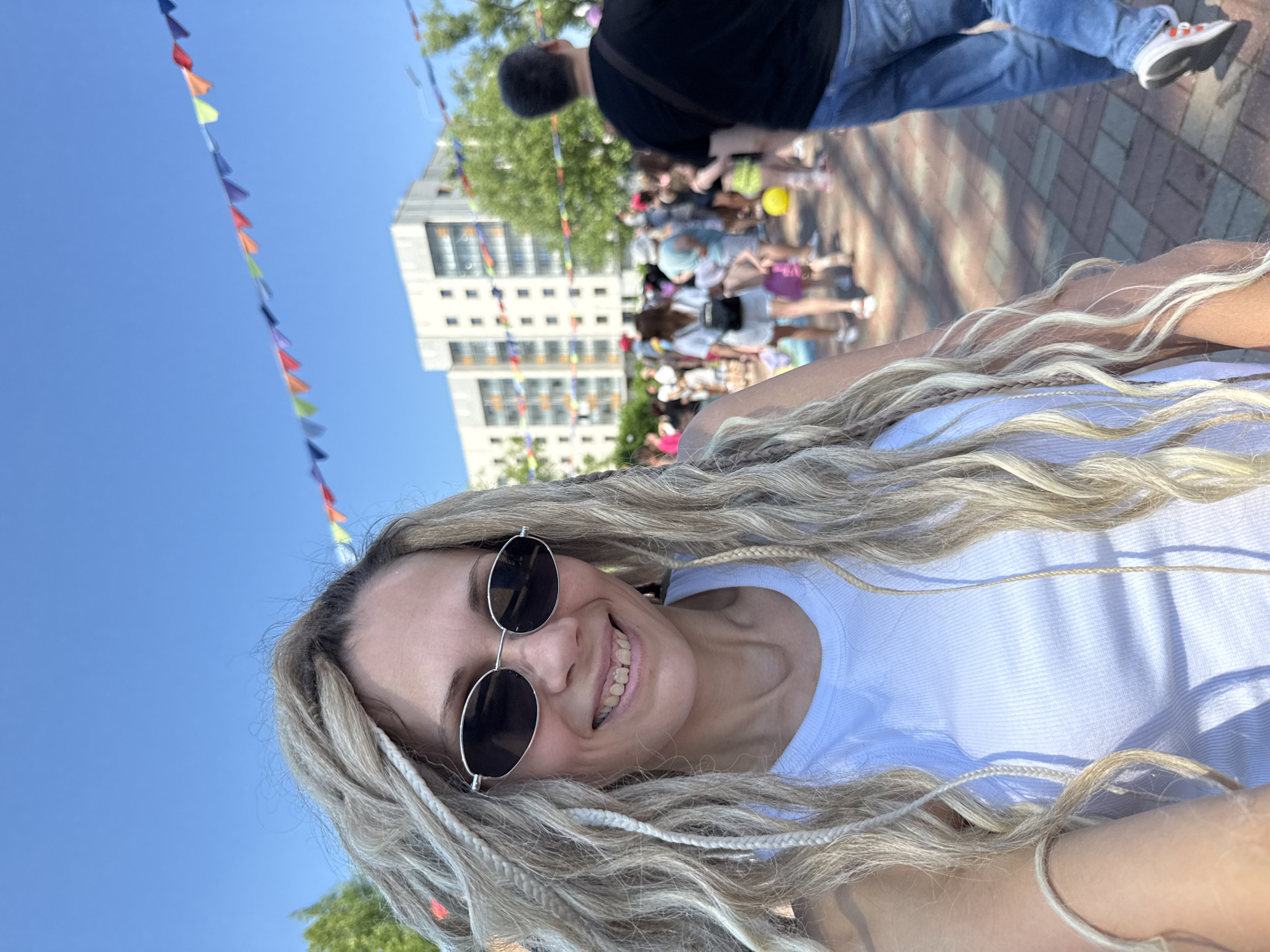 A medium shot shows a woman with blonde dreadlocks, wearing a white tank top and oval sunglasses, smiling at the camera. She is outdoors, with a blue sky overhead and a string of colorful pennant flags stretched across the upper portion of the frame. In the background, a modern, multi-story building with visible windows stands behind a crowd of people. Trees and more pennant flags are visible. The ground is paved with reddish-brown and grey bricks. The scene appears to be a public gathering or festival in the city of Zavolzhye, Russia.