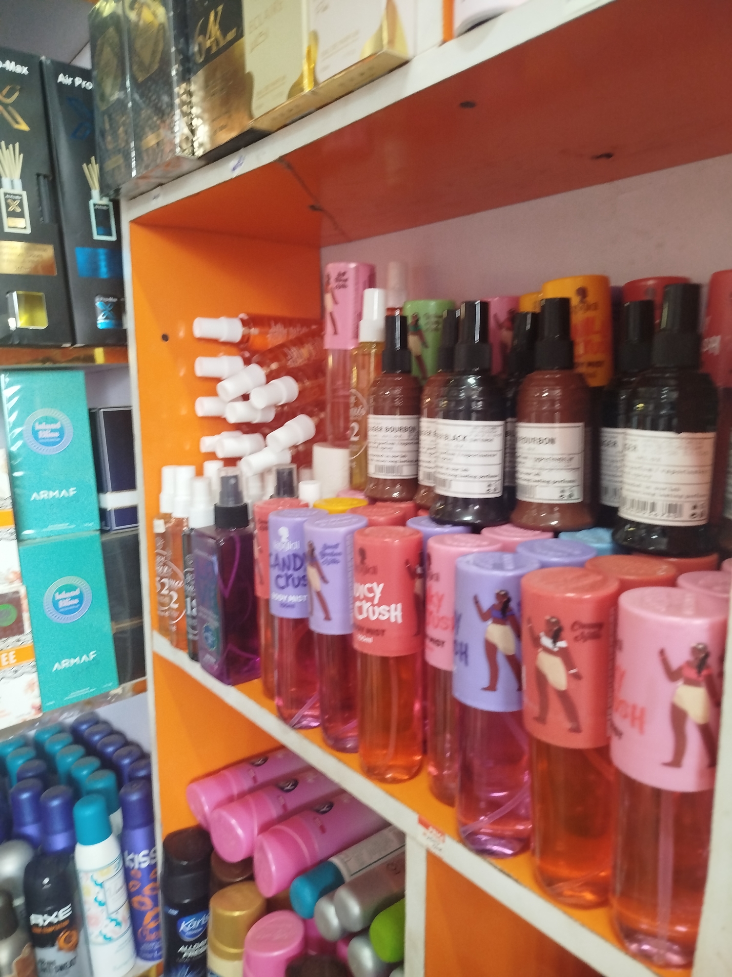 The image shows a close-up, slightly blurry view of shelves in a store in Ugbowo, Nigeria, stocked with various cosmetic and fragrance products. On the top shelf, several boxes of perfume are visible, including ones labeled "Air Pro" and "64K". The middle shelf is primarily filled with spray bottles of perfume and body mist, many with colorful labels featuring graphics of women. Brands like "Armaf," "Juicy Crush," and "Bourbon" are discernible. The bottom shelf displays an assortment of aerosol cans, likely deodorants or body sprays, with labels such as "AXE" and "Karl". The shelves themselves are made of white material with orange accents, suggesting a retail environment. The lighting is somewhat dim, with reflections visible on the product surfaces.