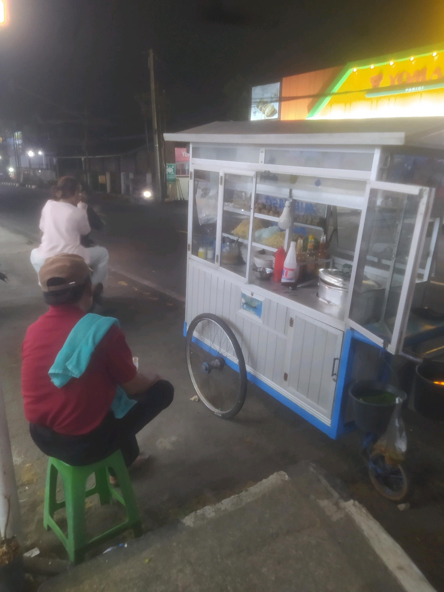 The image depicts a street scene at night in Desa Karangbenda, Indonesia. In the foreground, a man wearing a red shirt and a brown cap is seated on a green plastic stool. He has a light blue towel draped over his left shoulder. Beside him, a food cart with a white and blue facade is parked. The cart has glass display cases showing food items and bottles of condiments. A bicycle wheel is attached to the side of the cart. In the background, a person is riding a motorcycle. The surrounding environment appears to be a paved street with a sidewalk. A lit sign with yellow and green elements is visible above the food cart.