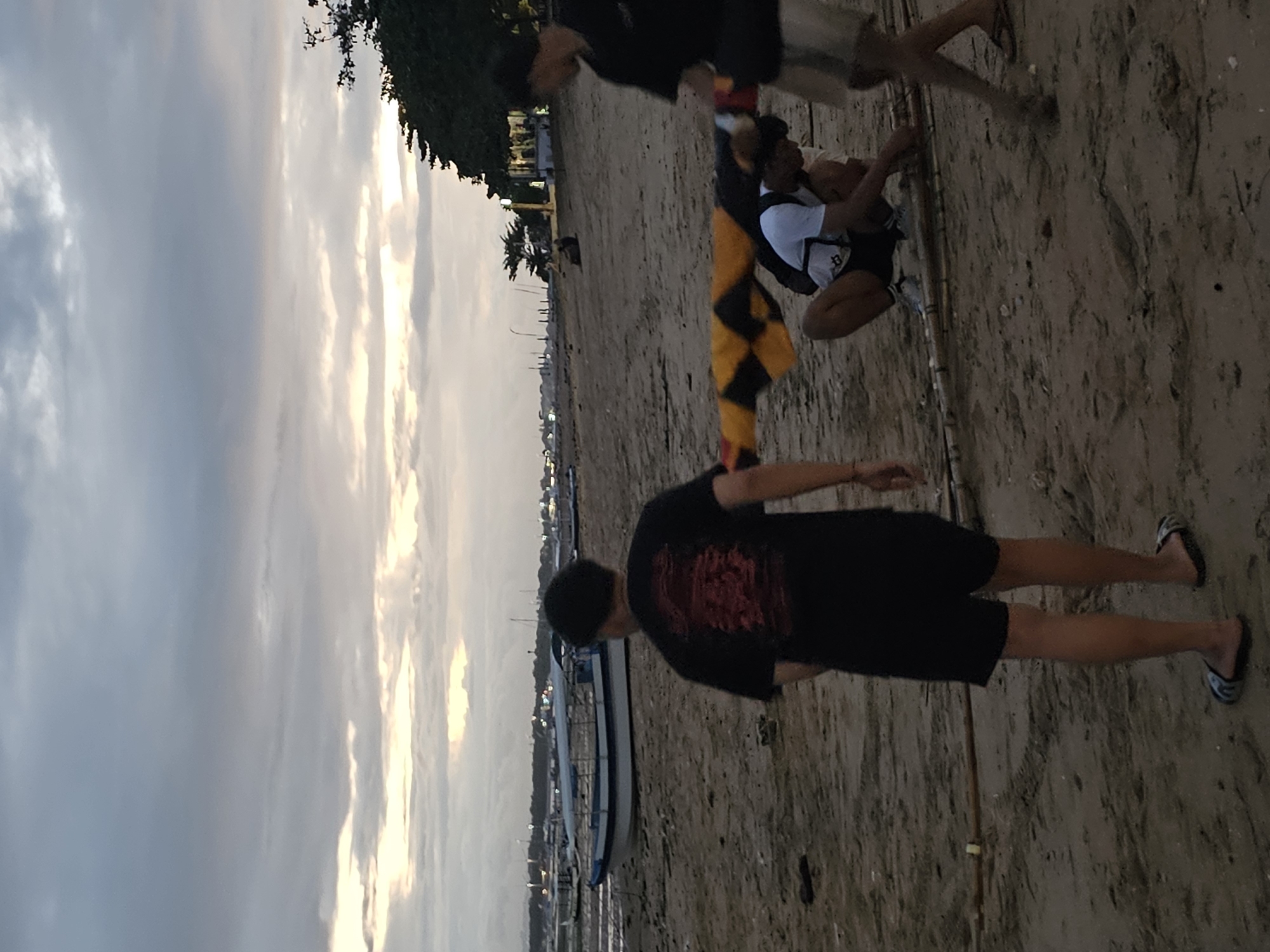 The image shows a group of people on a beach in Denpasar, Indonesia. The sky is cloudy. In the foreground, a person wearing a black t-shirt and black shorts is standing on the sand. Behind them, there is a boat pulled onto the beach. Further back, other people are visible, one of whom is wearing a yellow and black checkered garment.