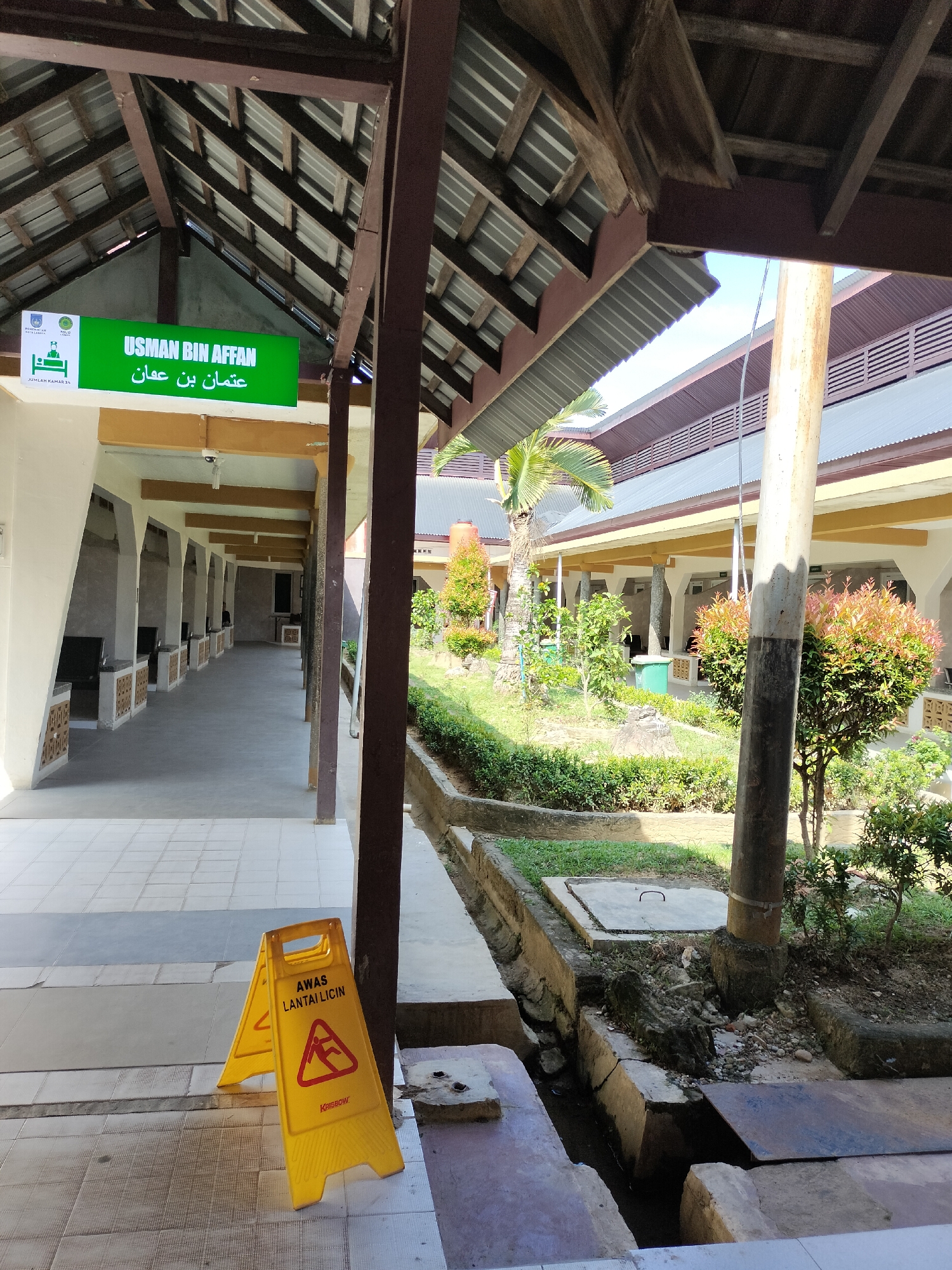 The image depicts a covered walkway with an adjacent garden area. The walkway features a tiled floor and a roof supported by dark brown beams and columns. To the left, a sign with Arabic and Latin script reads "USMAN BIN AFFAN" in green lettering, with accompanying logos. A yellow "wet floor" sign is visible in the foreground, indicating a slippery surface with a caution symbol of a person falling. Beyond the walkway, a well-maintained garden with various plants, shrubs, and a palm tree is present. In the background, buildings with tiled roofs and white walls are visible, suggesting an institutional setting. The overall impression is one of a functional and somewhat dated public space, likely part of a school, hospital, or religious institution. The location is identified as Langsa, Indonesia.