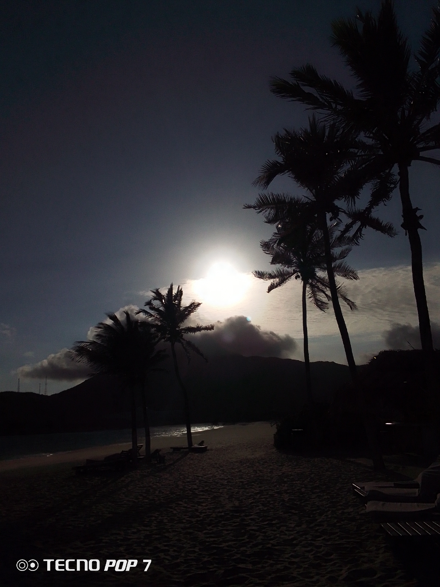 The image shows a beach scene in Pedro González, Venezuela. In the foreground, there is a sandy beach with beach chairs. Palm trees are silhouetted against the sun, which is obscured by clouds. In the background, a mountain range is visible. The TECNO POP 7 watermark is visible in the lower left corner.