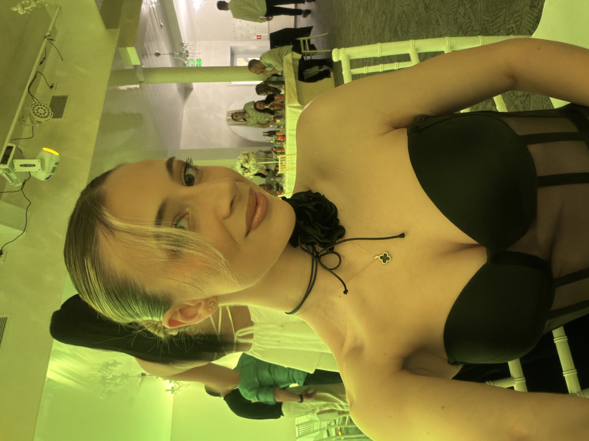 A woman is centered in the frame, posing for a selfie-style photograph. She has blonde hair styled to the side with a dark black sleeveless dress. She is wearing a black necklace with a clover shaped pendant and black flower detail.
The background reveals an indoor setting, likely a restaurant or event hall, given the tables and chairs set up for a gathering. People are seated around the tables in the distance, suggesting a social event. The location appears to be within Plovdiv, Bulgaria, but further specifics are not discernible from the image.