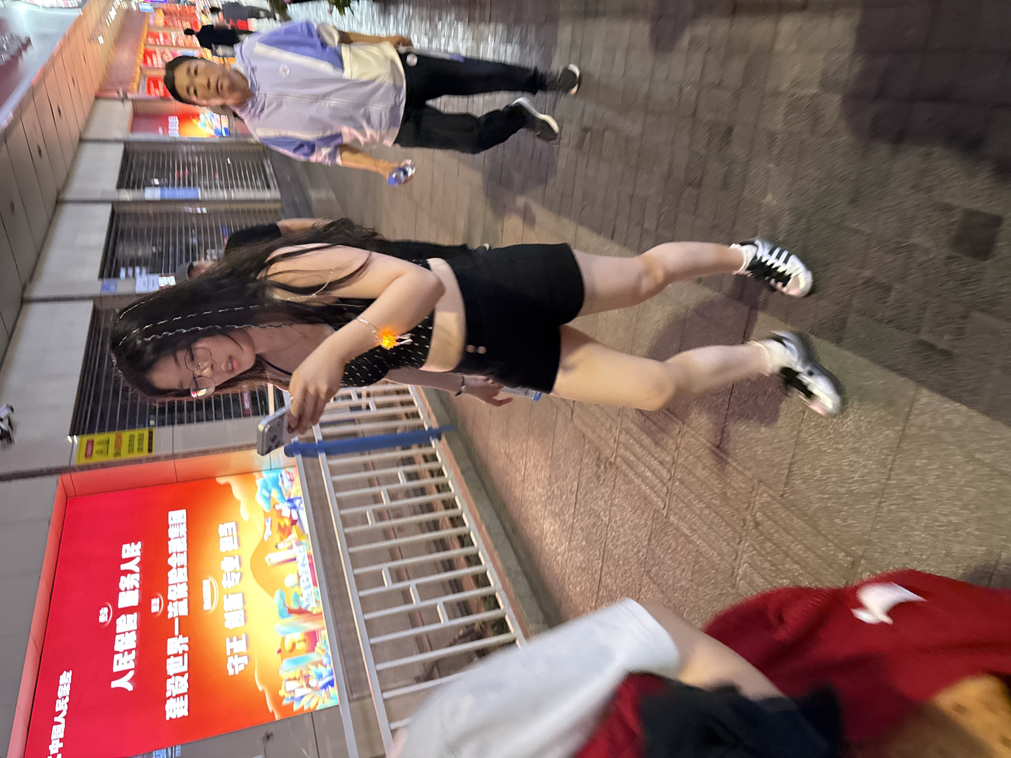 A woman is looking at her phone in Yuzhong District, China. She is wearing glasses, a black top with white polka dots, black shorts, and black and white striped tennis shoes. She has long, dark hair. Another person is out of focus and partly visible in the bottom of the photo, wearing a red sweater. A man wearing a light-colored jacket and black pants is walking behind her. The background includes a building with closed storefronts, a red, lighted sign, and a metal railing. The sign is in Chinese. The location is a city street.