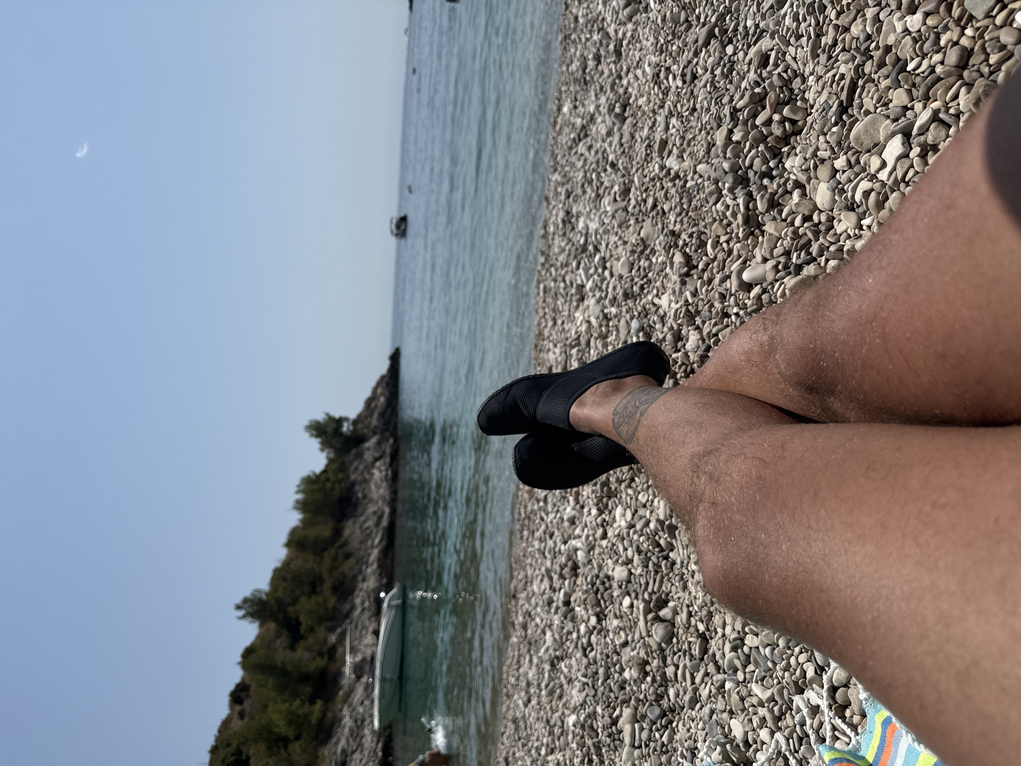 The image depicts a person's legs and feet, clad in black water shoes, crossed while resting on a pebbled beach. In the background, the clear turquoise waters of the Adriatic Sea meet a rocky coastline dotted with green foliage. A white motorboat is anchored in the shallow water near the shore, and other boats are visible further out at sea. The sky is a clear, pale blue with a faint crescent moon visible. The scene is set in a coastal area, likely Hvar town, Croatia, known for its beautiful beaches and clear waters. The overall impression is one of relaxation and enjoyment of a sunny day at the seaside.
