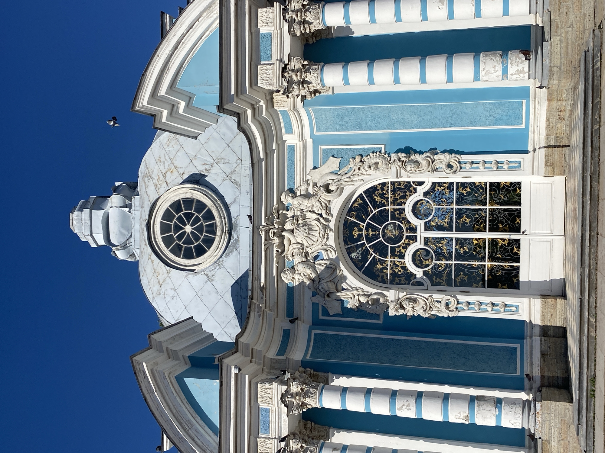 This image shows a frontal view of the Church of the Transfiguration in the Catherine Park, Pushkin, Russia. The church is painted in a bright blue and white color scheme, with ornate white stucco decorations and columns. A large, arched window with intricate gold tracery is prominently featured in the center of the facade, above a white double door. Above the window, a circular window with a sunburst pattern is visible, and higher still, a domed roof with a small cupola. The sky is a clear, deep blue. A pigeon is captured in flight in the upper right portion of the frame, and several pigeons are perched on ledges and columns. The foreground shows a stone staircase leading up to the entrance, with a checkered tile pattern visible on the ground. The overall impression is one of architectural grandeur under a bright, sunny sky.