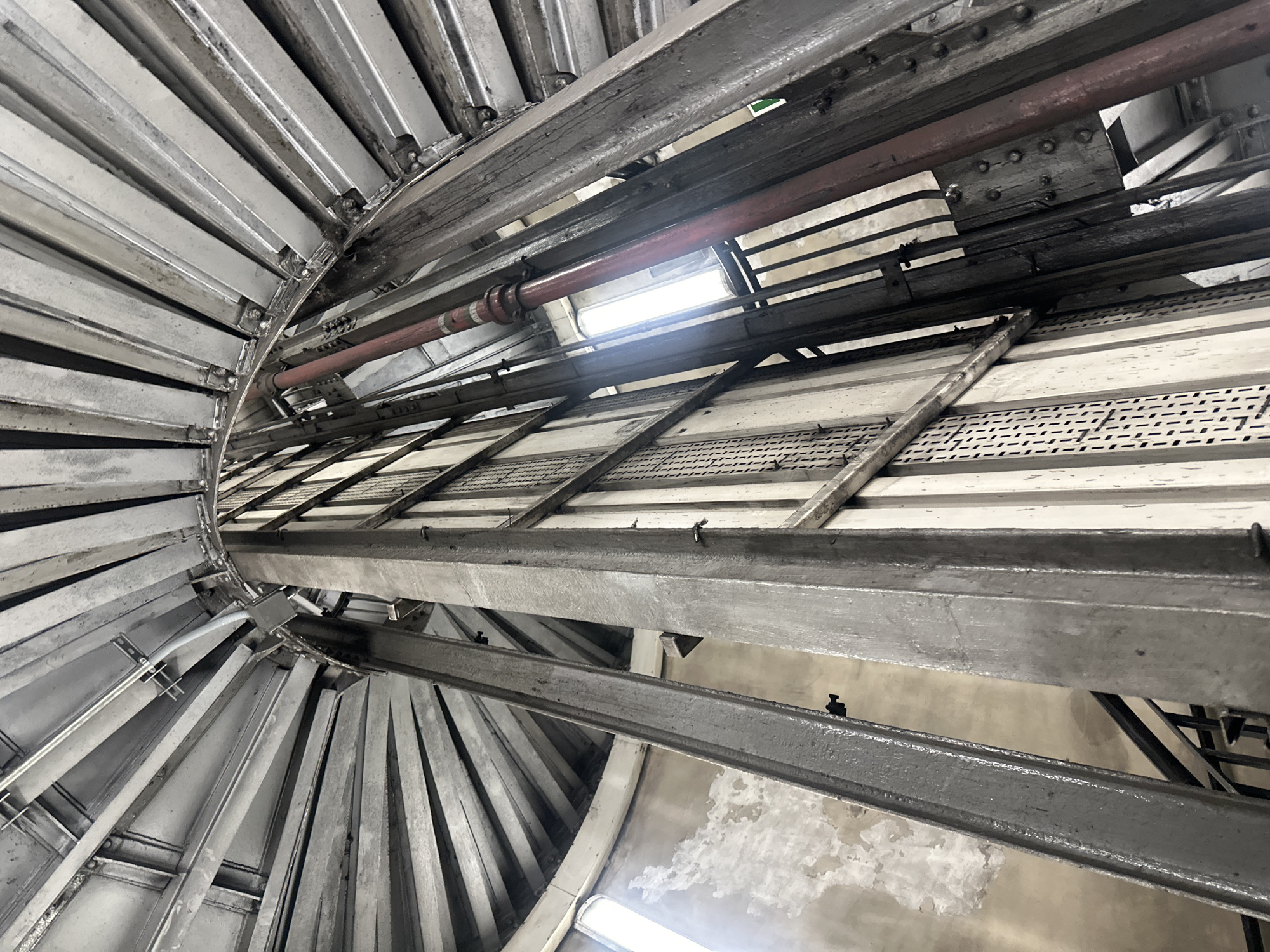 The image depicts the interior of a structure, likely a tower or a large industrial building, with a prominent spiral staircase on the left side. The staircase is constructed of metal and ascends upwards. To the right of the staircase, there's a complex arrangement of metal beams, supports, and a red pipe. The ceiling or upper section of the structure has a perforated metal grid, and there are openings allowing light to enter, creating bright spots in the scene. The walls visible between the metal structures appear to be made of concrete or a similar material, and there are signs of wear and possible water damage. This photograph could be located at the ArcelorMittal Orbit sculpture in London, United Kingdom, which offers a unique interior perspective with industrial-style construction.