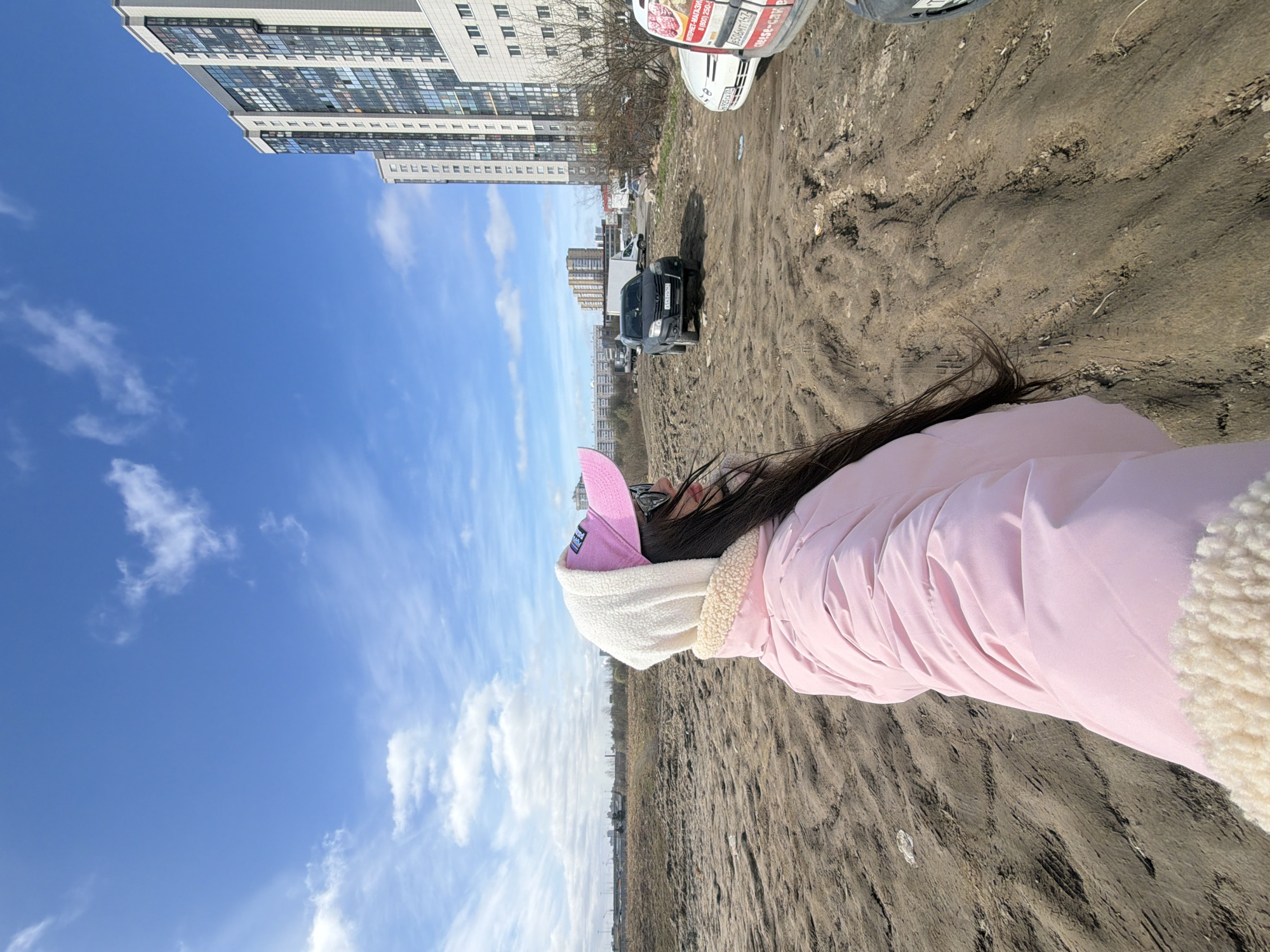 The image depicts a woman wearing a pink jacket and hat standing on a muddy, sandy area near a city landscape. In the background, there are buildings characteristic of urban development. The sky is blue with scattered clouds. Based on the architecture and overall environment, the location is likely Saint Petersburg, Russia.