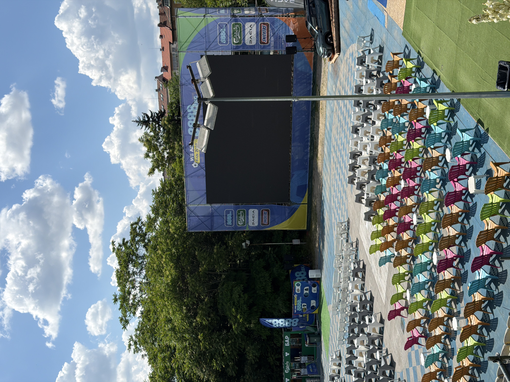 This image depicts an outdoor cinema or event space. In the background, a large screen is set up with various sponsors' logos, including 8888.bg, Carlsberg, Dacia, and Snickers. The screen is surrounded by blue and green banners. A line of trees forms the backdrop, with some buildings visible beyond the trees. In the foreground, rows of colorful chairs, mostly Adirondack-style, are arranged in neat rows on a blue checkered ground. There are also white chairs and some benches placed further back. To the left, a bar area with green stalls and flags is visible. A dark-colored SUV is parked on the right side of the frame, positioned on wooden logs. The scene is set on a bright, sunny day with a partly cloudy sky. This outdoor venue is located in Sofia, Bulgaria, and appears to be a fan zone, possibly for a sporting event or a summer cinema.
