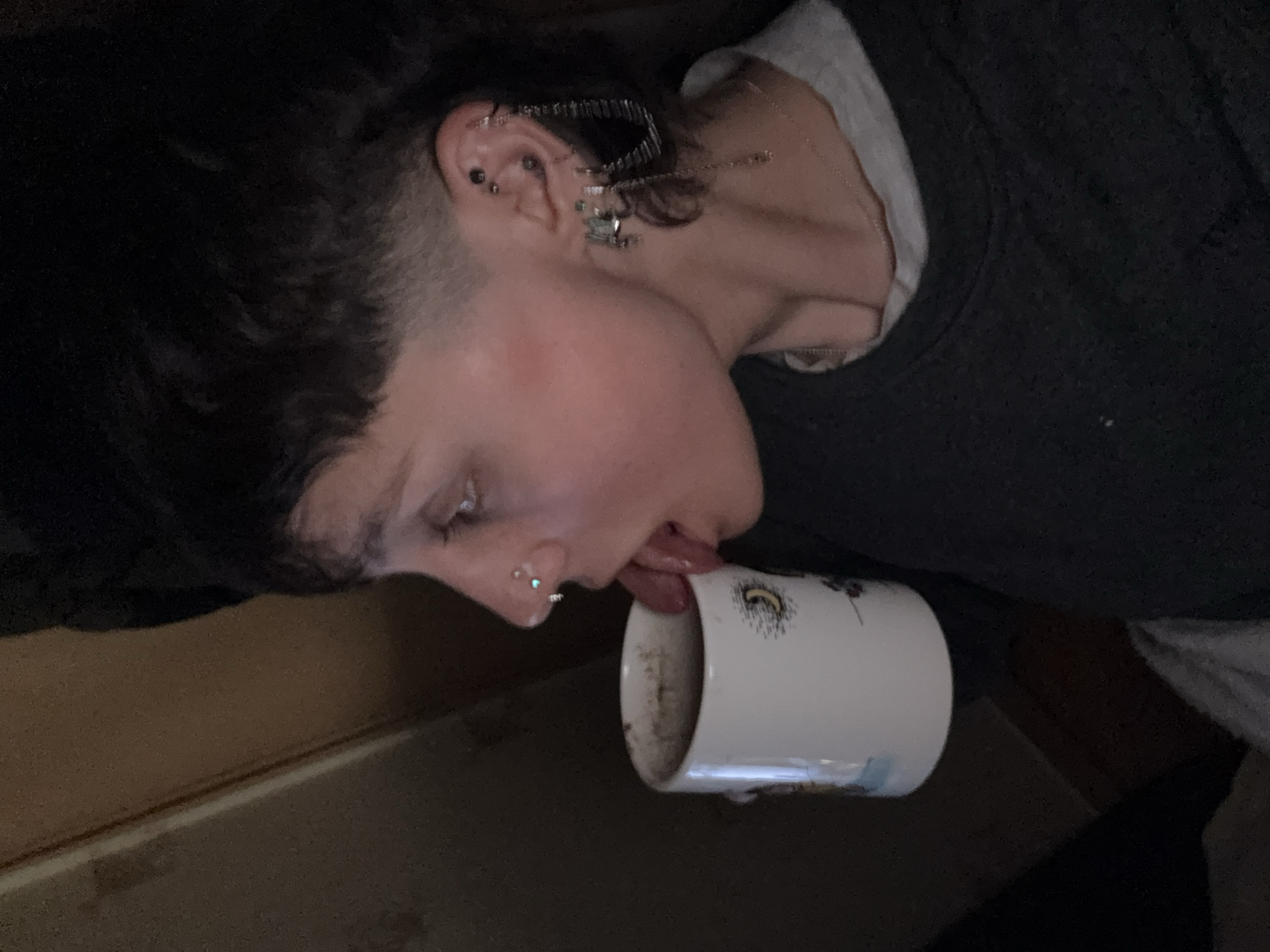 A person with multiple piercings in their ear and a nose piercing is licking the inside of a white mug. The person has short, dark hair with a shaved section on the side. The mug appears to be mostly empty and has a design on its side. The background is blurry, but includes a dark surface and a lighter, possibly wooden, surface. There is no indication this image was taken in the city of Moscow, Russia.