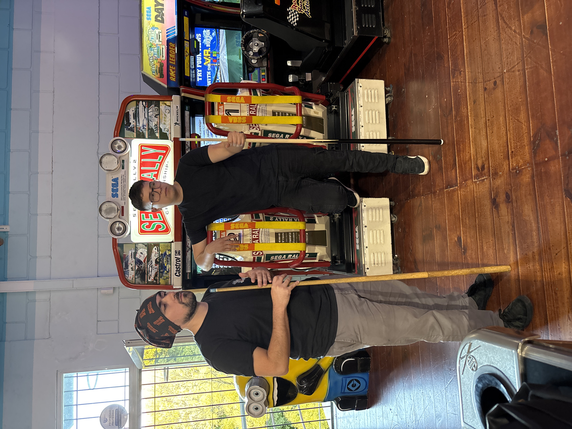 Two men hold pool cues horizontally in front of two racing arcade games with "SEGA" and "RALLY 2" on the marquees. The man on the left wears a bandana and a t-shirt with gray pants. The man on the right wears glasses and a t-shirt with black pants. A minion figurine sits next to the left arcade game. The room has wooden floors and blue-and-white walls. A window is on the left with outdoor vegetation visible. This is likely a recreation center or arcade game room. The text on the window reads "PROPIEDAD PROTEGIDA". The location appears to be Villa General Belgrano, Argentina based on the text on the bandanna of the person on the left: "ROYAL ENFIELD".