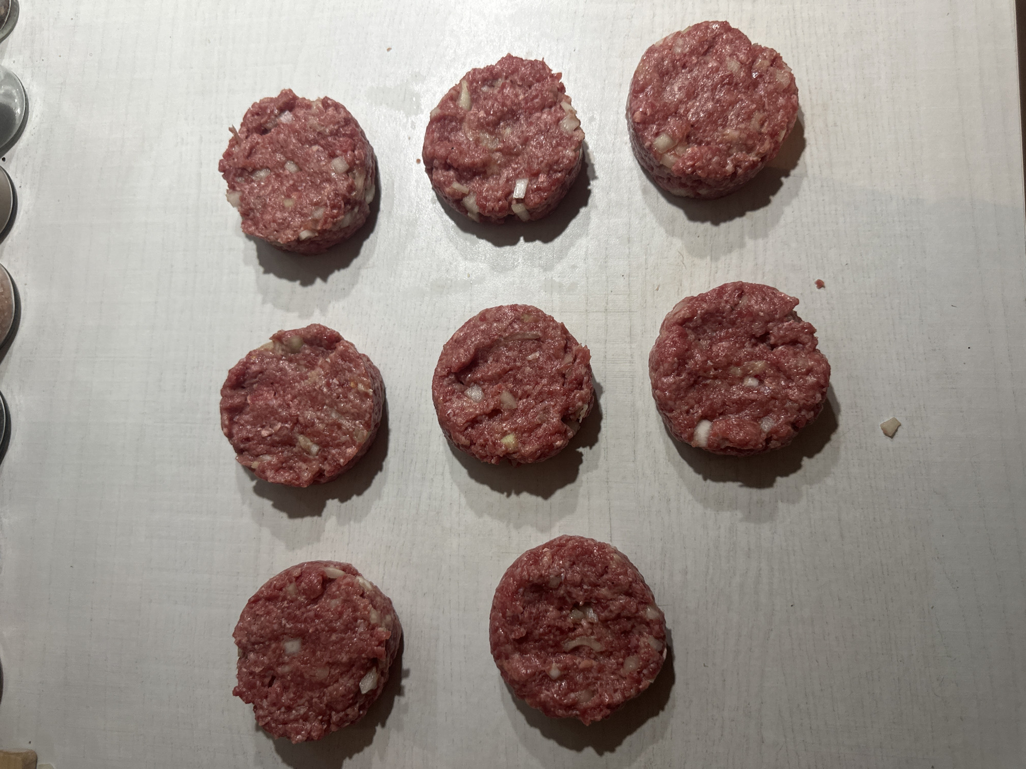 Nine raw ground meat patties are presented on a light, textured surface, likely a kitchen countertop or cutting board. There are nine patties in total, arranged in an informal grid-like pattern across the frame. Each patty is round, composed of reddish-brown ground meat, and clearly shows small, irregular pieces of white, indicating the presence of chopped onions mixed into the meat. The patties appear freshly prepared and uniform in size.
The surface beneath the patties is light grey or off-white with subtle lines and imperfections, suggesting a worn or textured material. Shadows cast by the patties indicate overhead lighting, adding depth to the scene. On the far left edge of the image, parts of metallic objects, possibly measuring spoons or small kitchen containers, are partially visible, reinforcing the kitchen setting.
The scene depicts an intermediate stage of food preparation, with the patties ready for cooking. No people are visible, and there is no discernible text within the image. The overall impression is one of simple, home-style meal preparation.