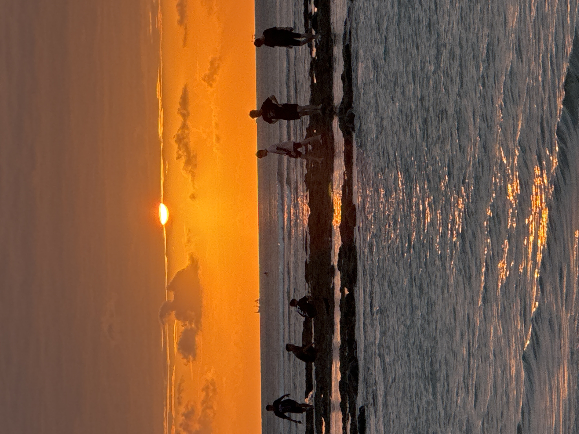 The image depicts a sunset at a rocky beach in Kuta Selatan, Indonesia. The sky is dominated by vibrant orange and yellow hues from the setting sun, with scattered clouds. The sun is partially obscured by clouds, casting a warm glow over the scene. In the foreground, the ocean water reflects the sunset's colors, creating shimmering patterns on the surface.
Several individuals, silhouetted against the bright sky, are scattered across the rocky shore. On the left, a person is walking out of the water. Further along the rocks, three individuals are crouched or sitting. In the middle ground, a person stands facing left, and to their right, another person stands with hands on their hips. On the far right, a person walks away from the camera.
In the distance, beyond the group on the rocks, a body of water stretches to the horizon. A ship is visible in the distance, near the horizon line. The overall scene captures a serene moment at sunset, with people enjoying the coastal environment.