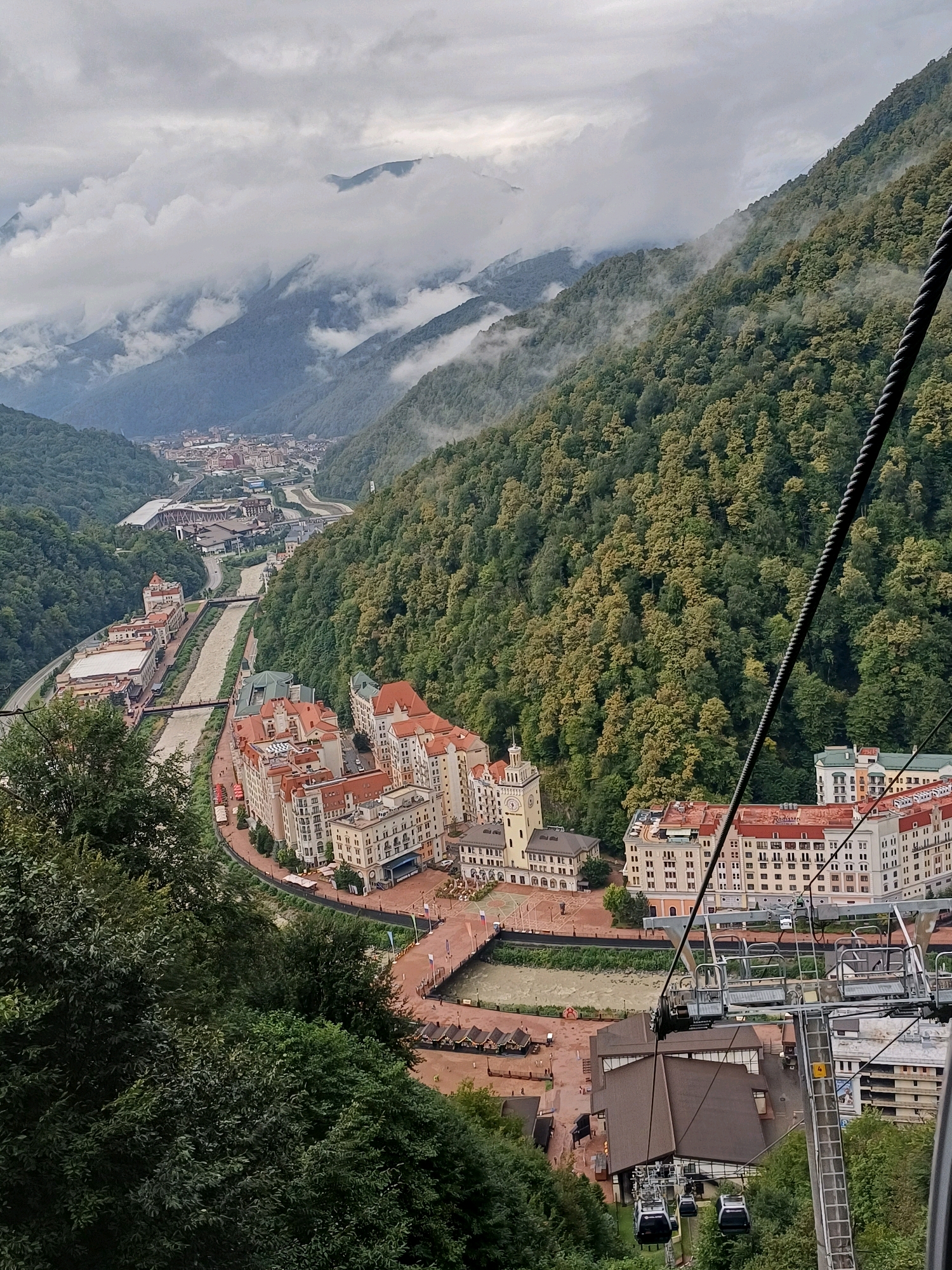The image shows a panoramic view of the Esto-Sadok ski resort in Krasnodar Krai, Russia, from the perspective of a cable car. The resort is nestled in a valley surrounded by lush, forested mountains. A river runs through the valley, with modern buildings lining its banks. The buildings are multi-story structures with red-tiled roofs and light-colored facades, typical of European alpine architecture. A cable car system is visible in the foreground, with several cabins suspended from a thick cable. The mountains in the background are covered in dense green trees, with clouds partially obscuring the peaks, suggesting a misty or overcast day. The overall scene depicts a picturesque mountain resort town.