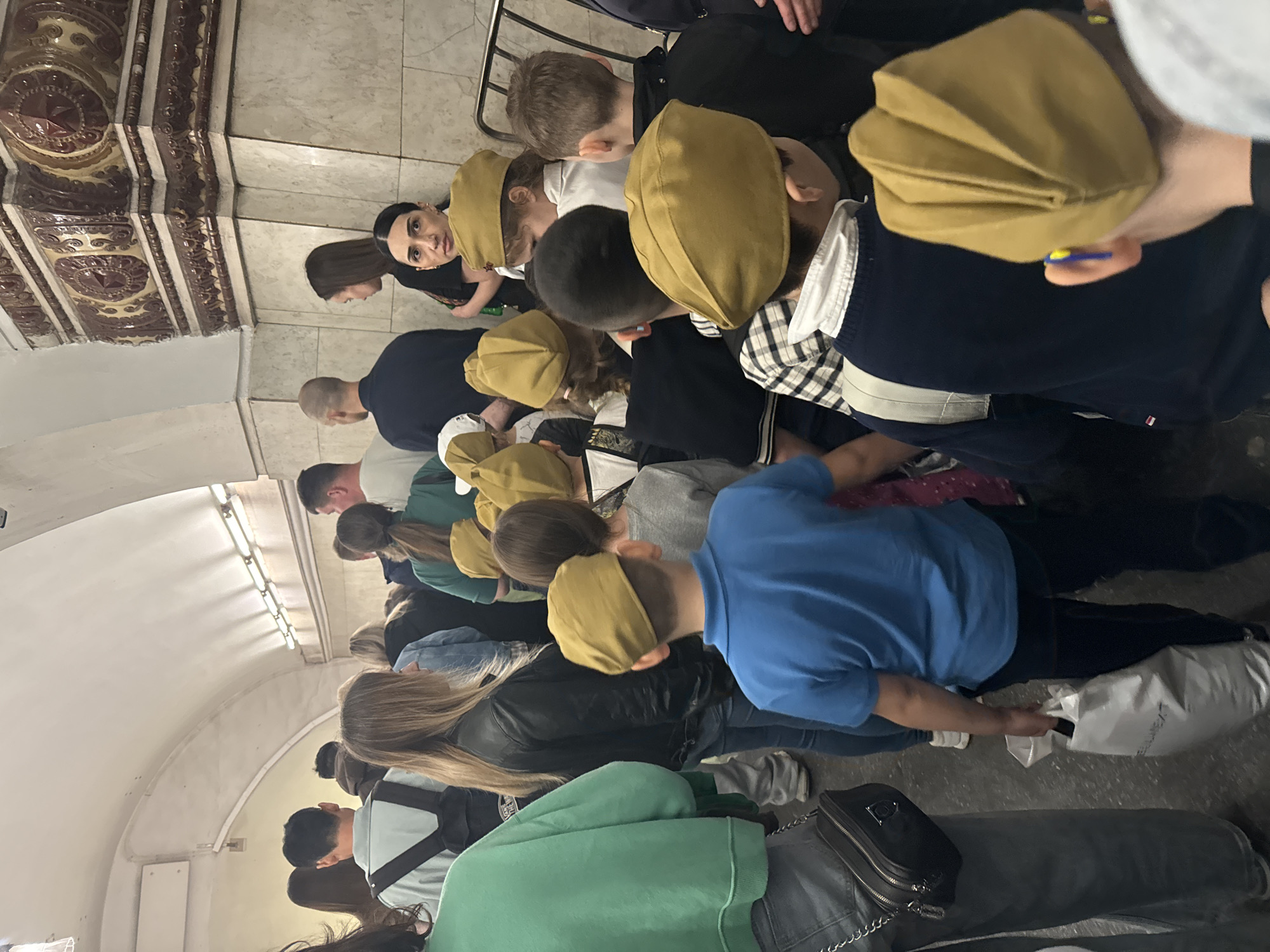 A group of people, many wearing khaki-colored hats, are gathered in what appears to be a subway station in Moscow, Russia. The station features decorative patterns on the walls and ceiling. The people are standing close together, some facing forward and some turned to the side. There are women with long hair, men with short hair, and several children in the group. Some people are wearing casual clothing like jeans, t-shirts, and jackets. One person in the foreground is wearing a green sweatshirt and a gray bag. Another individual is wearing a blue shirt and has a white shopping bag.