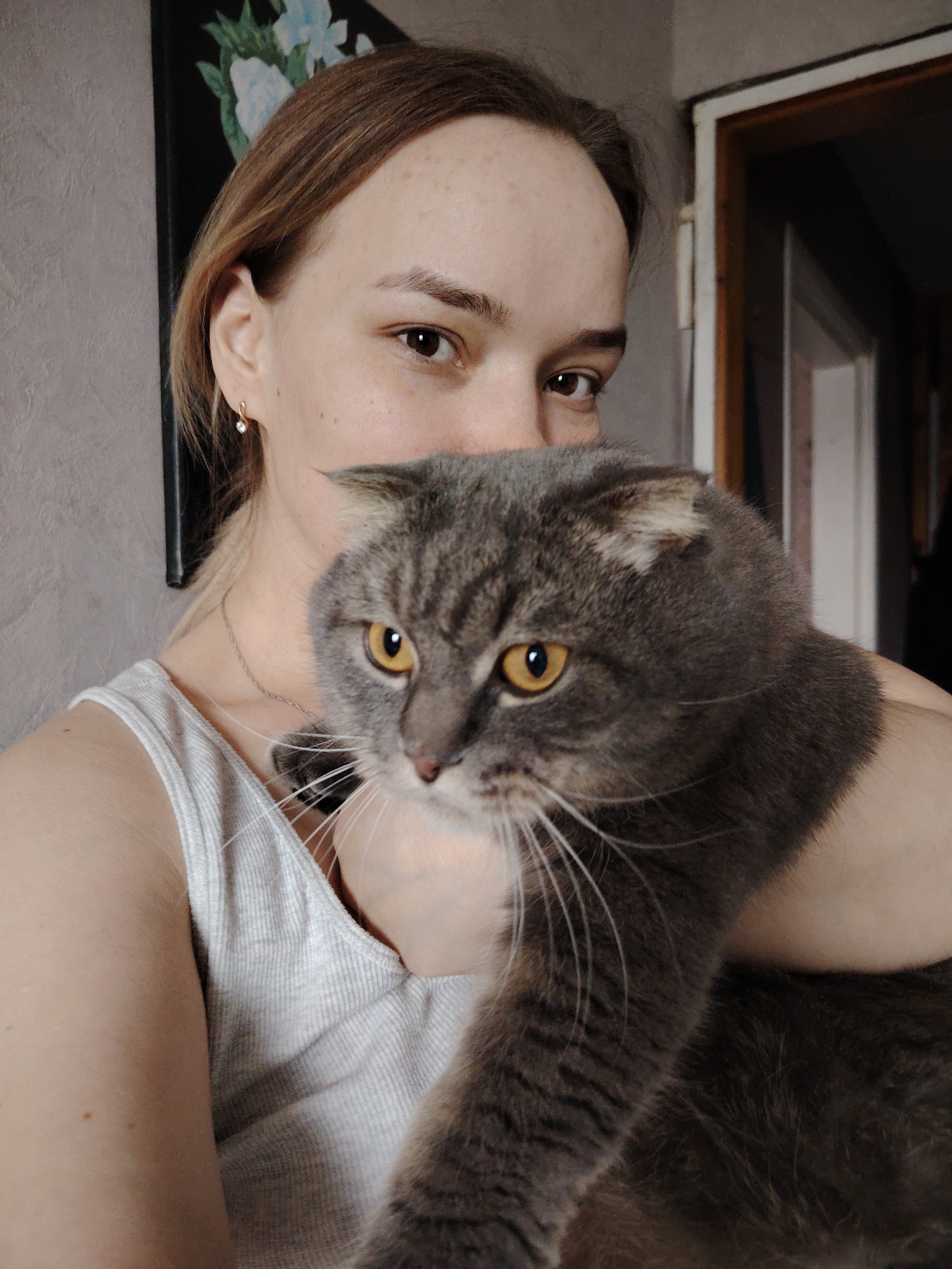 A woman is holding a Scottish Fold cat. The woman has fair skin, light brown hair pulled back, and is wearing a light gray tank top and a small gold earring. The cat is gray with orange eyes. Behind the woman is a gray wall with a dark framed picture containing flowers. To the right, there is an open doorway. It's unlikely this photo was taken in the city of Марковский, Russia; this information is likely a hallucination.