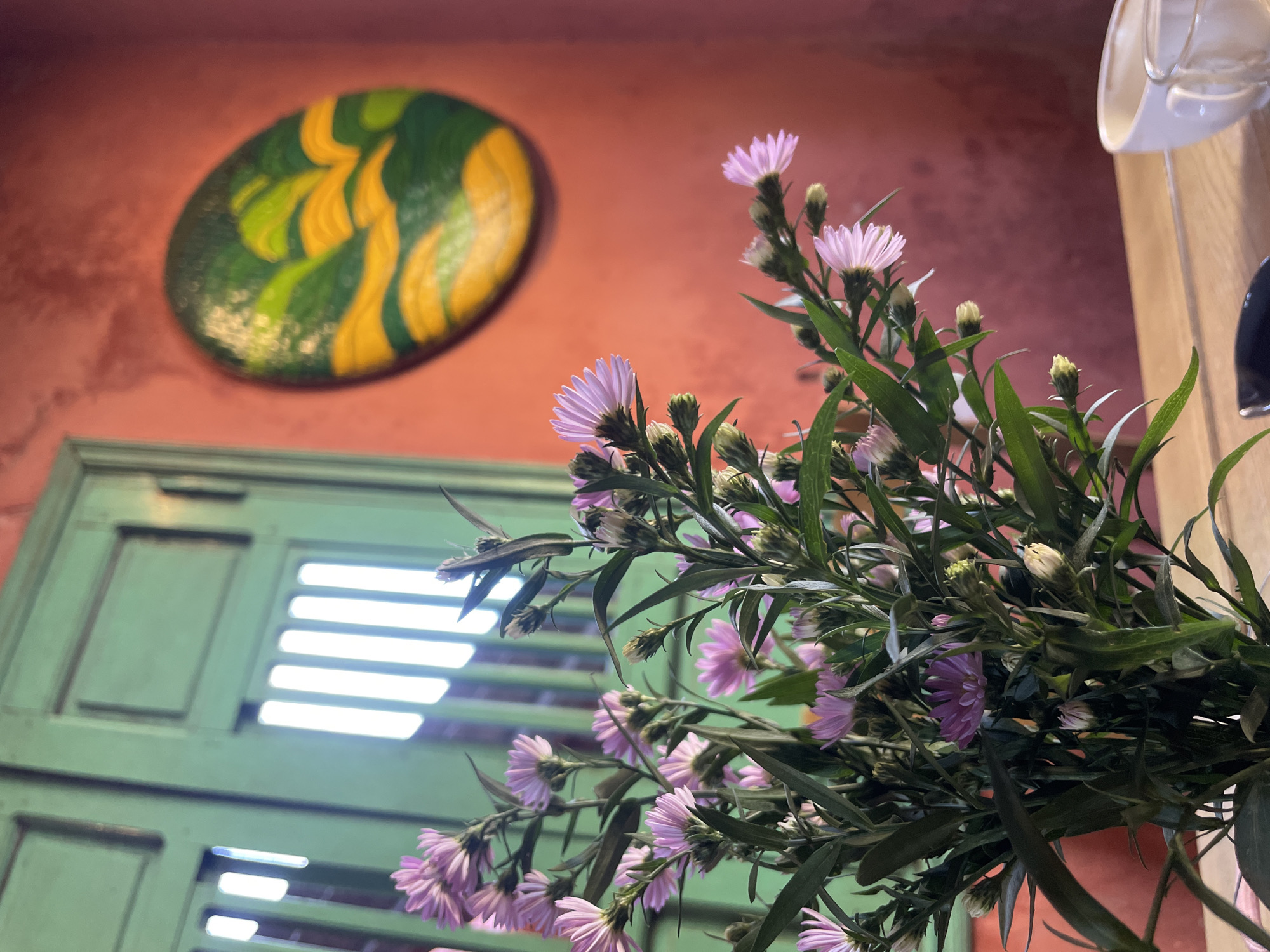The image depicts a floral arrangement indoors. The flowers are predominantly lavender with green foliage, arranged in a vase that is not fully visible. Behind the flowers, a green shuttered window is slightly ajar, allowing bright light to stream in. The walls are painted in a textured reddish-orange hue, and a decorative oval painting with green and yellow designs is visible on the wall. The scene is set in Hội An Ward, Vietnam.