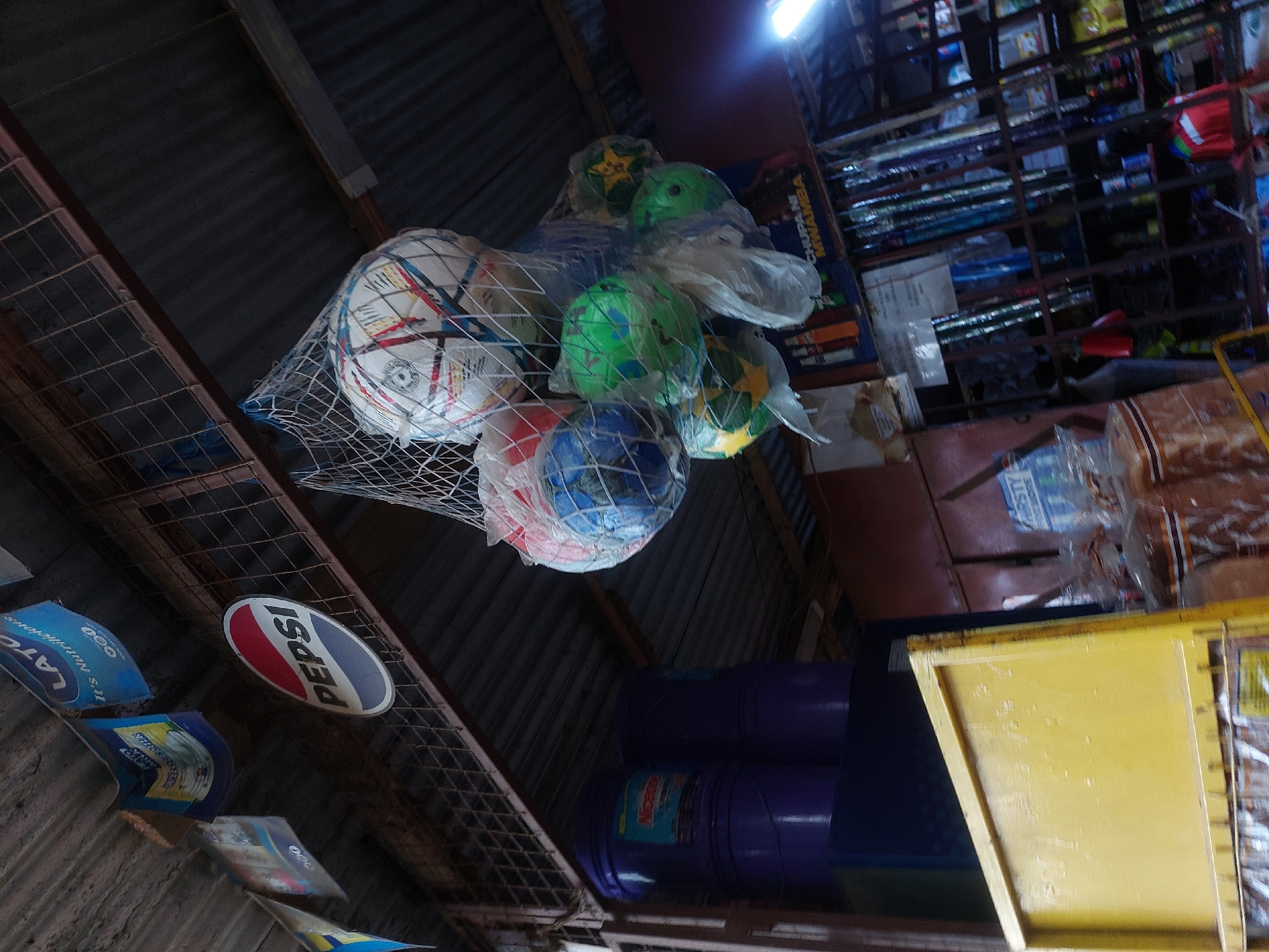 The image shows the interior of a small shop in Dar es-Salaam, Tanzania. The shop has a corrugated metal roof and a metal grid frame visible. A net filled with inflated soccer balls hangs from the ceiling. Some balls are white with geometric patterns, while others are green and blue. Several advertising signs for "Lato" and "Pepsi" are attached to the walls. The shop also features shelves stocked with various goods in the background, as well as stacks of purple buckets and a yellow counter.