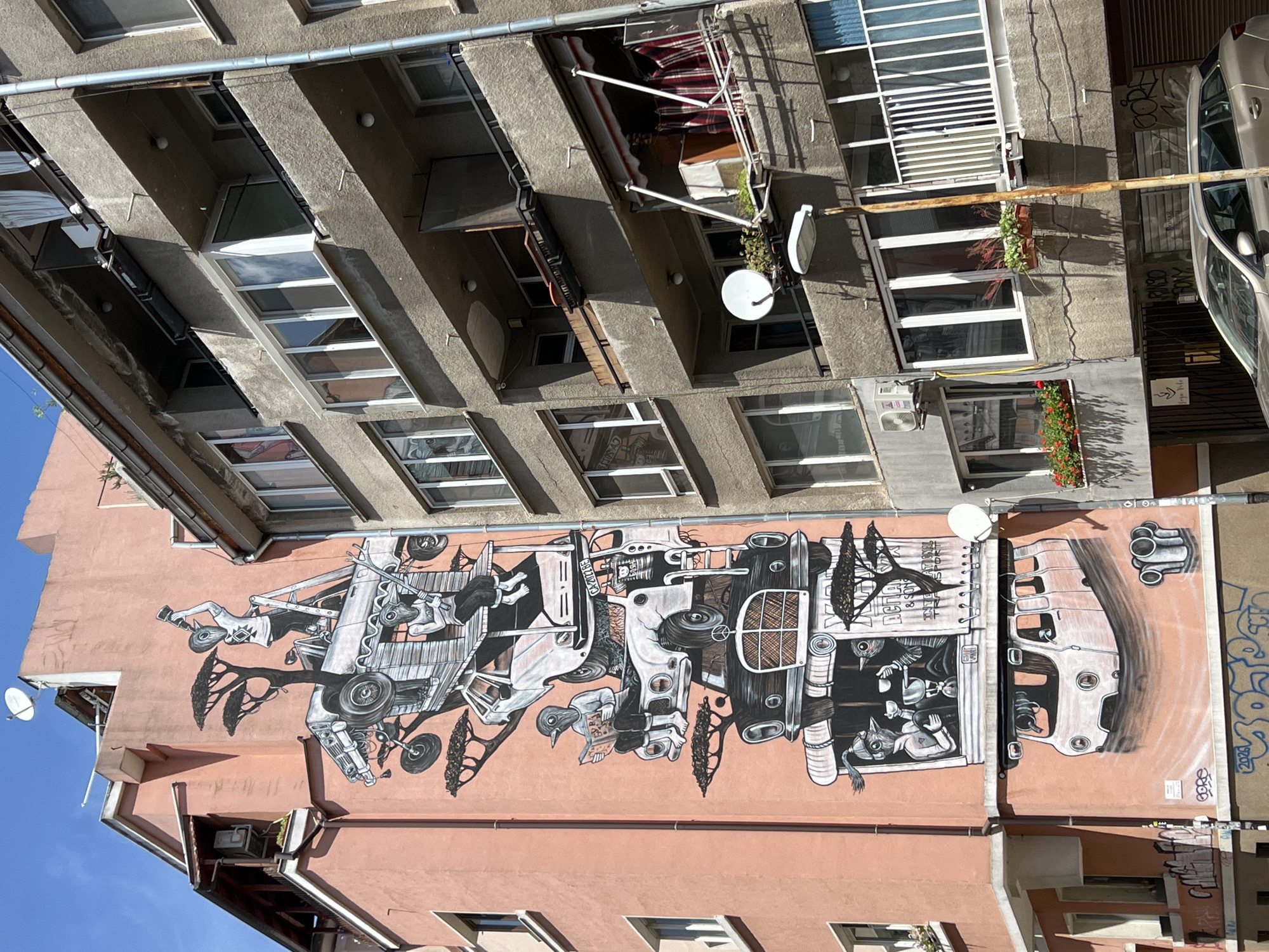The image displays a building in Sofia, Bulgaria, featuring a prominent mural on its pink-colored facade. The mural depicts a surreal arrangement of stacked vehicles and figures. At the top, a figure resembling a bird-like creature wearing a mask stands on a ladder, holding a telescope and pointing it upwards. Below this, another vehicle is stacked, with a similar bird-like figure sitting inside. Further down, a figure is seated in an open-top vehicle, reading a book. Another vehicle, a black vintage car, forms the base of this stacked structure. At the bottom of the mural, a white van appears to be levitating above a swirling vortex. The mural is intricate, with stylized trees and various details within the vehicles.
Adjacent to the pink building is a gray concrete apartment building with multiple balconies. Some balconies have potted plants and clotheslines with laundry. A satellite dish is visible on one of the balconies, along with an outdoor light fixture and a lamp post. The gray building has numerous windows, some with visible reflections of the sky and surrounding buildings. The scene is captured under a clear blue sky. Graffiti is present on the lower parts of both buildings, including on a parked silver station wagon in the foreground. The overall impression is an urban environment with a striking piece of street art.