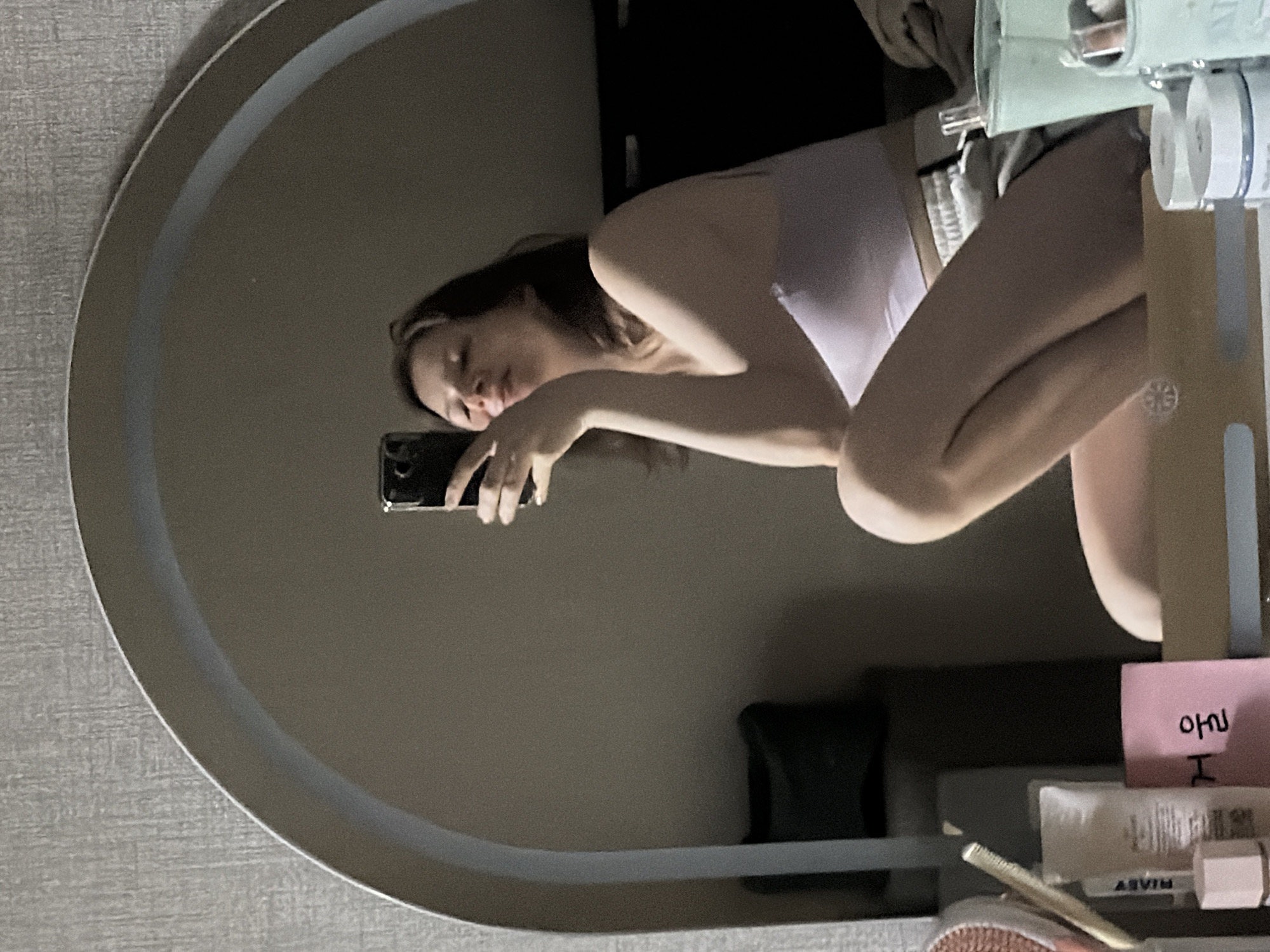 A young woman is taking a selfie in a mirror, reflecting her upper body and legs as she sits with her knees drawn up. She is wearing a light purple camisole and grey shorts. She holds a black smartphone in her right hand, with her left hand resting on her chin. The mirror is large and arched, framed by a light-colored border, and positioned against a textured grey wall. In the foreground, on a surface below the mirror, various cosmetic items are visible, including a pink notepad with Korean writing, a tube of cream, and a small white jar. The lighting is dim, suggesting an indoor setting, likely a bedroom or dressing area. The reflection shows a portion of a bed and a dark object that could be a pillow in the background. There are no indications of the city of Dongducheon-si, South Korea.