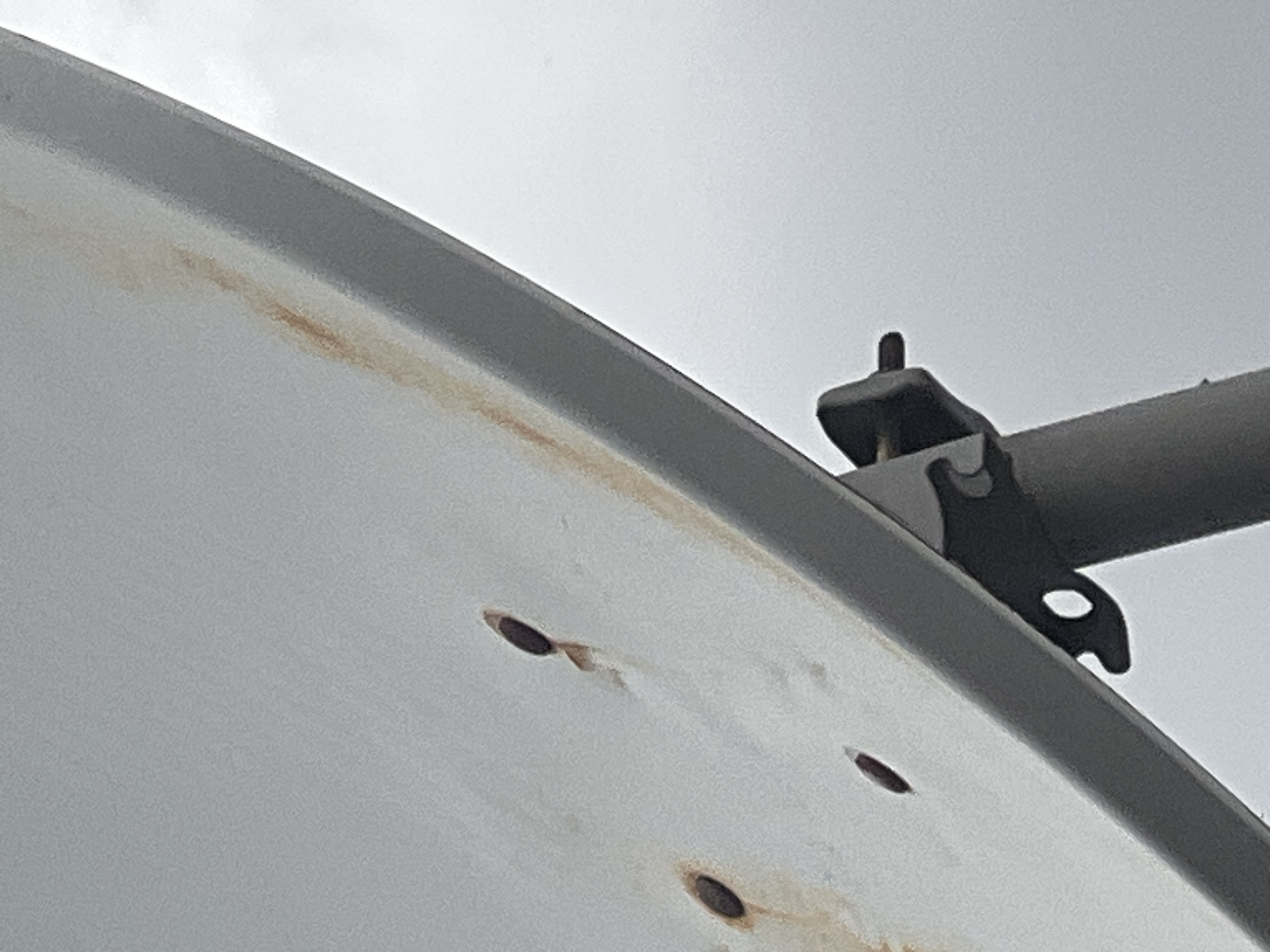 The image shows a close-up of a white satellite dish, tilted upwards against a cloudy sky. The dish has several small, dark, oval-shaped markings that appear to be rust spots. There is also a streak of rust running down the side of the dish. A dark grey metal bracket is attached to the dish, connecting it to a pole that is visible at the bottom of the image. The overall impression is of a weathered object, likely exposed to the elements. The scene is set in Jalingo, Nigeria, an environment characterized by its tropical climate, which can contribute to the rusting of exposed metal objects.