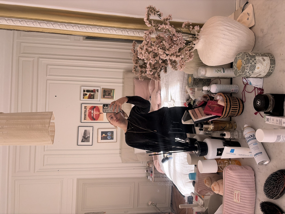 The image displays a mirror selfie of a woman taken in a bedroom, identified as an apartment in Paris, France, by the presence of characteristic Haussmannian boiseries (decorative wall paneling) and a tall, paneled white door on the left.
The individual, a fair-skinned woman with light hair pulled back, is seen from the chest up, wearing a dark long-sleeved top and dark trousers. She holds a smartphone with her raised left hand, taking the photograph. Her head is tilted to the left, and her lips are pursed. She wears two layered necklaces and rings on her left hand.
The foreground consists of a marble or light-colored vanity surface cluttered with numerous personal care products, cosmetics, and decorative items. These include two dark hairbrushes, a pink quilted Clarins pouch, various bottles and tubes (La Roche-Posay, Klorane, EtiaXil visible), a black reed diffuser, a small woven basket containing makeup palettes, a Chanel container, and a patterned Byredo candle box. A textured white ceramic vase containing dried light pink flowers is prominent on the right side of the vanity.
Reflected in the mirror behind the woman is a bedroom area. A large bed with a light-colored fabric headboard, white bedding, and several light pink throw pillows occupies the center. Above the bed, a gallery wall features seven framed artworks of varying sizes, including a "PLAYBOY" poster with red lips, abstract pieces, and a black and white photograph. A light-colored fabric pendant lamp hangs from the high ceiling. A golden-framed mirror or architectural element is visible on the far right of the reflection.