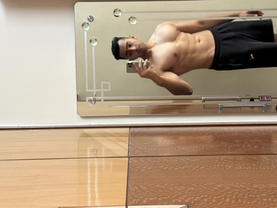 The image depicts an indoor scene where a young adult male of South Asian appearance takes a mirror selfie. The male subject, shirtless and wearing black athletic shorts with a white Puma logo and a green side stripe, stands in front of a rectangular mirror with rounded corners. He holds a silver-colored smartphone in his right hand, angled to capture his reflection. A gold-colored bangle is present on his left wrist. The mirror features an etched geometric border design and clear, circular decorative elements. To the left of the mirror, a multi-panel wooden wardrobe or cabinet is visible, consisting of lighter, polished wood on its upper sections and darker, textured wood on its lower sections. The wall behind the mirror is light-colored. Reflections in the mirror also show parts of vertical, brushed metal handles, indicating an adjacent door or cabinet. The setting is a private residential interior, consistent with a bedroom or dressing area, located in Kolkata, India. The precise address or neighborhood within Kolkata cannot be ascertained from the visual evidence.
