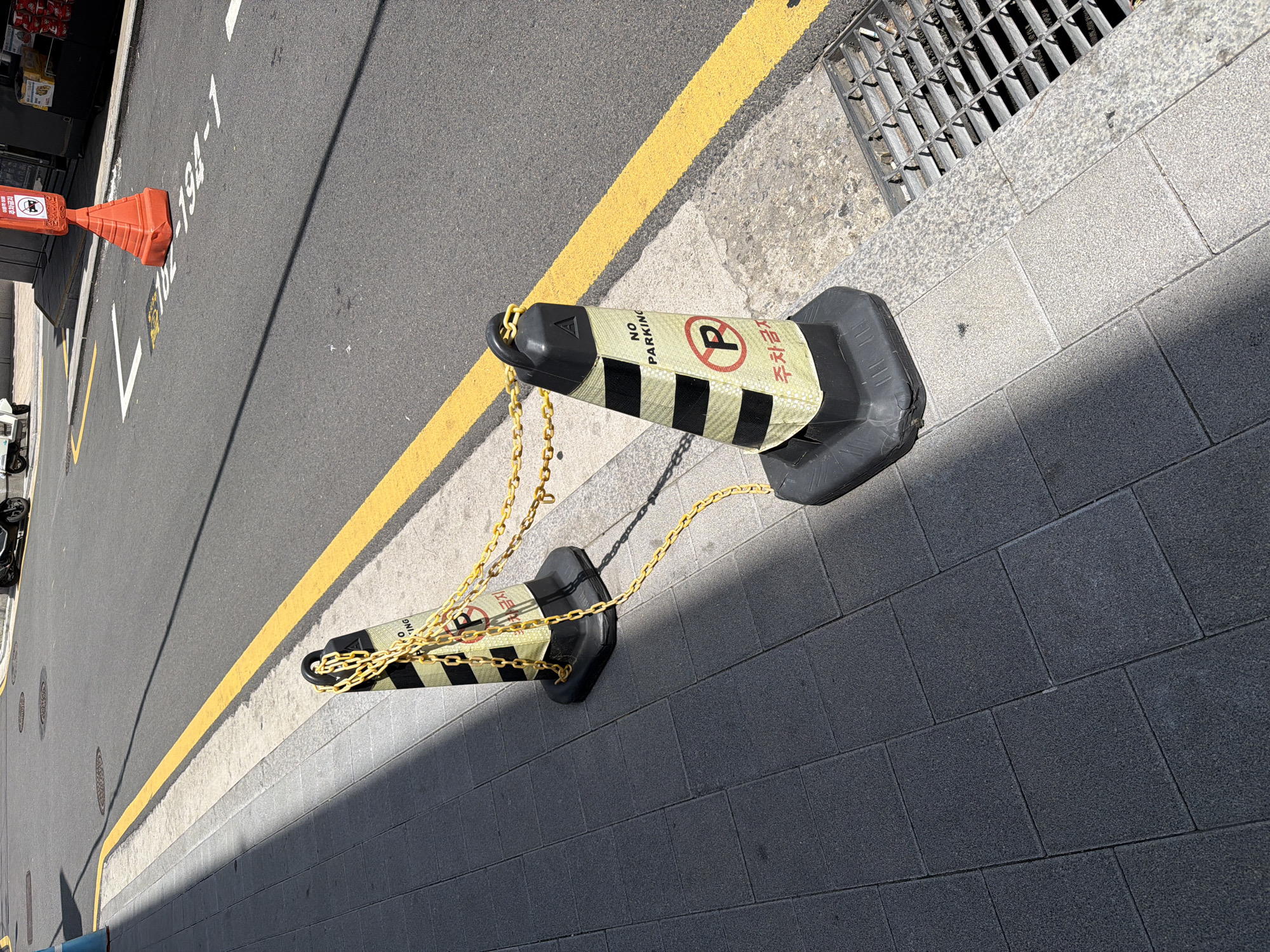 The image shows a street scene in Seoul, South Korea. Two black-and-yellow traffic cones are chained together on the sidewalk next to a curb painted with a solid yellow line. The cones bear the text "NO PARKING" in English and "주차금지" (pronounced "jucha geumji") in Korean, which also means "no parking". An orange traffic cone is visible on the roadway in the distance. A metal grate drain cover is visible near the top right of the image. The sidewalk is made of gray square paving stones. The roadway is asphalt.
